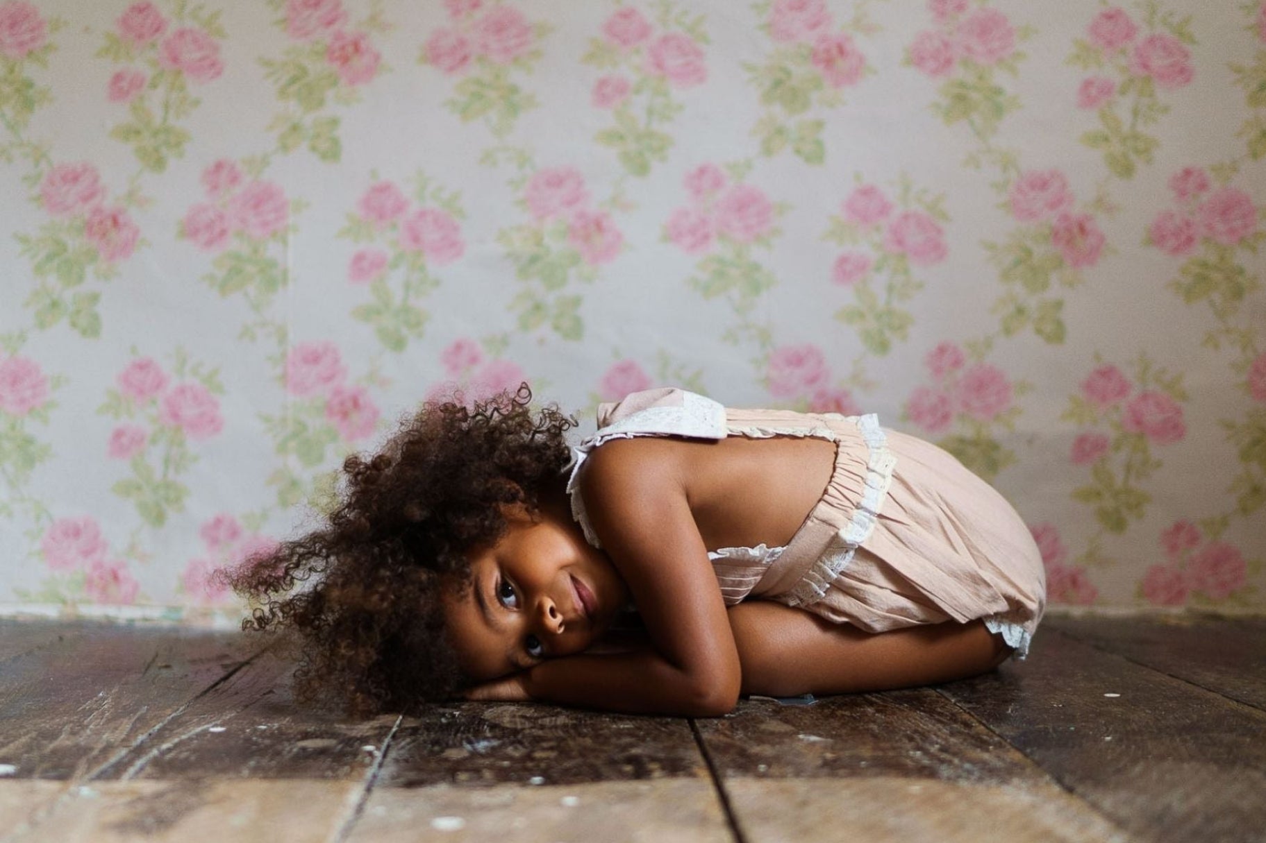 A young child lying on a wooden floor wearing a powder pink romper made from organic cotton, set against a vintage floral wallpaper backdrop, embodying slow, natural, and ethical children's clothing.