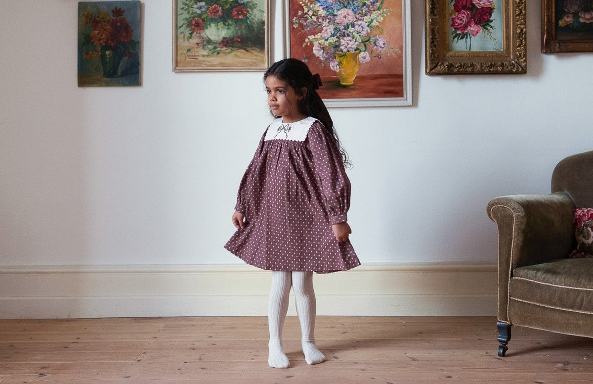 Young girl wearing a vintage style burgundy dress with a white collar and long sleeves in a room decorated with floral paintings and an antique armchair
