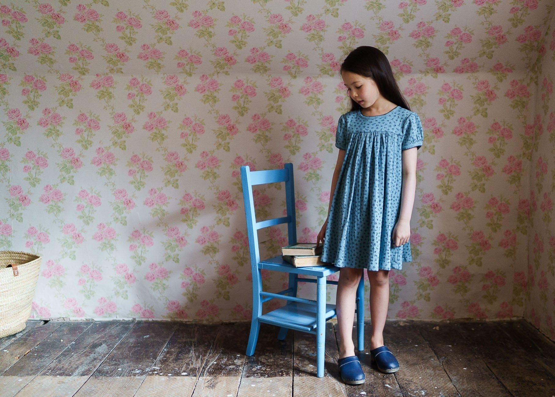 Young girl wearing a blue floral organic cotton dress standing beside a vintage-style blue wooden chair with a stack of books, in a room with floral wallpaper and wooden floor.