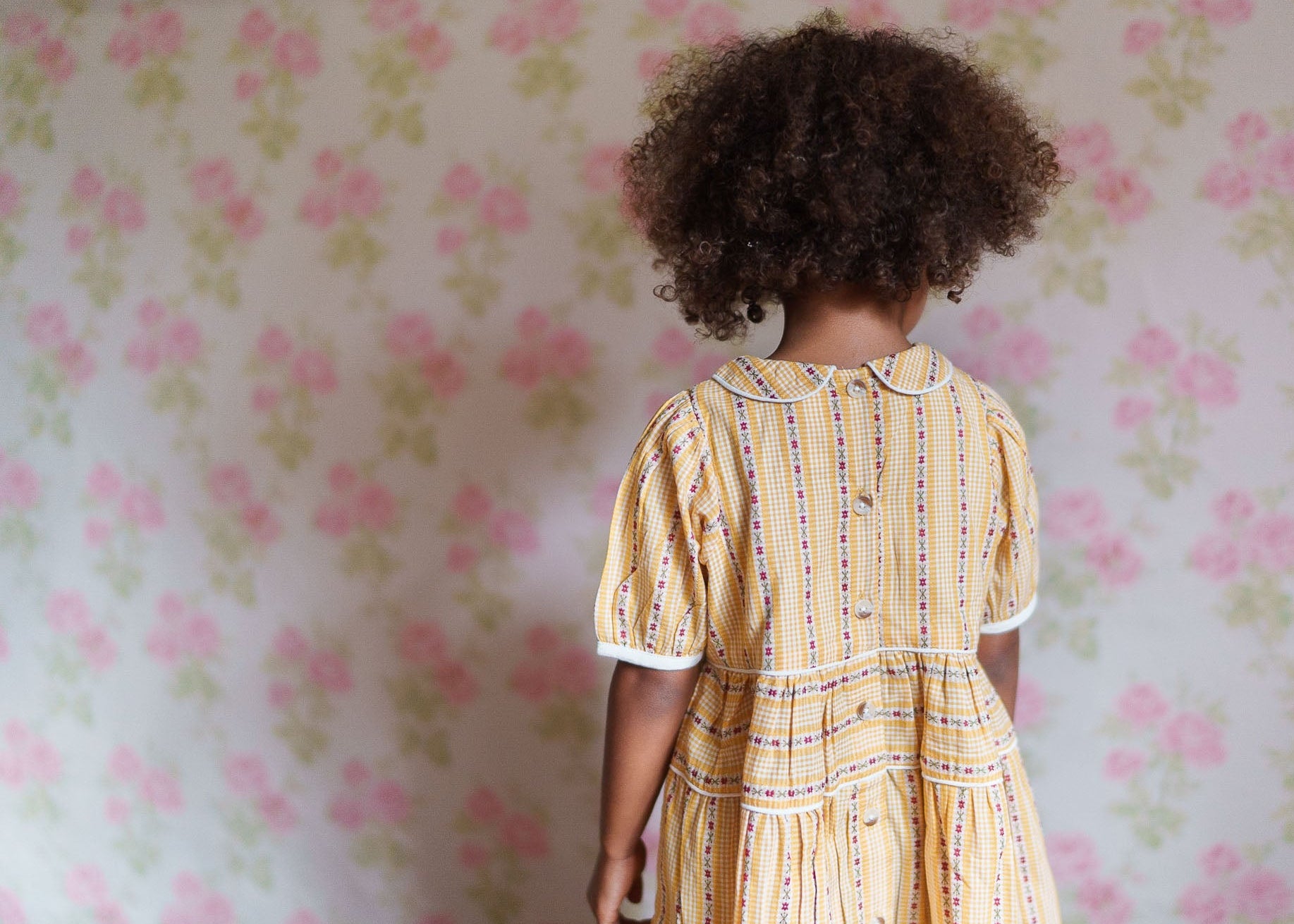 Young child wearing a yellow floral organic cotton dress with a vintage style, standing against a soft pink floral wallpaper background.