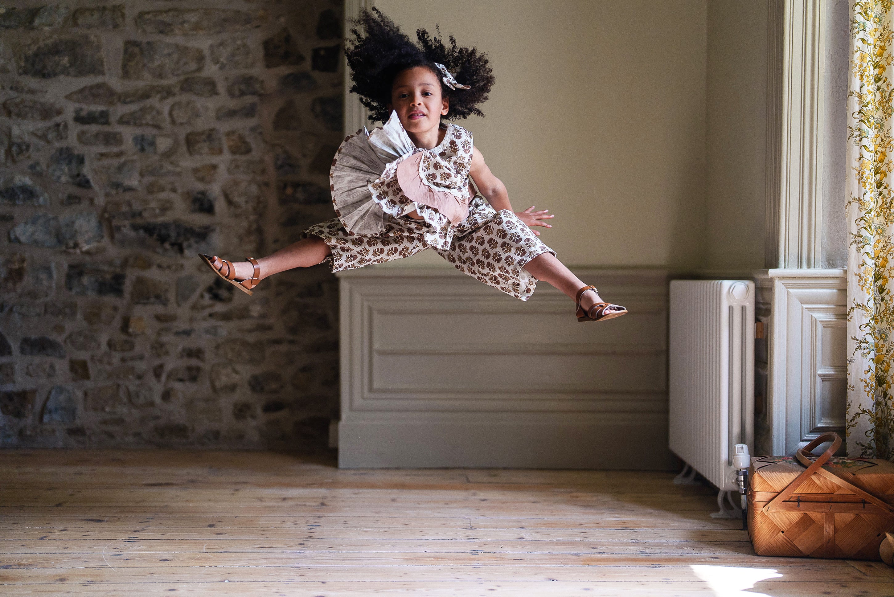 A joyful child wearing a pink and white floral vintage-style girls blouse and matching bottoms, made from organic cotton in a sunlit room with rustic wooden floors.