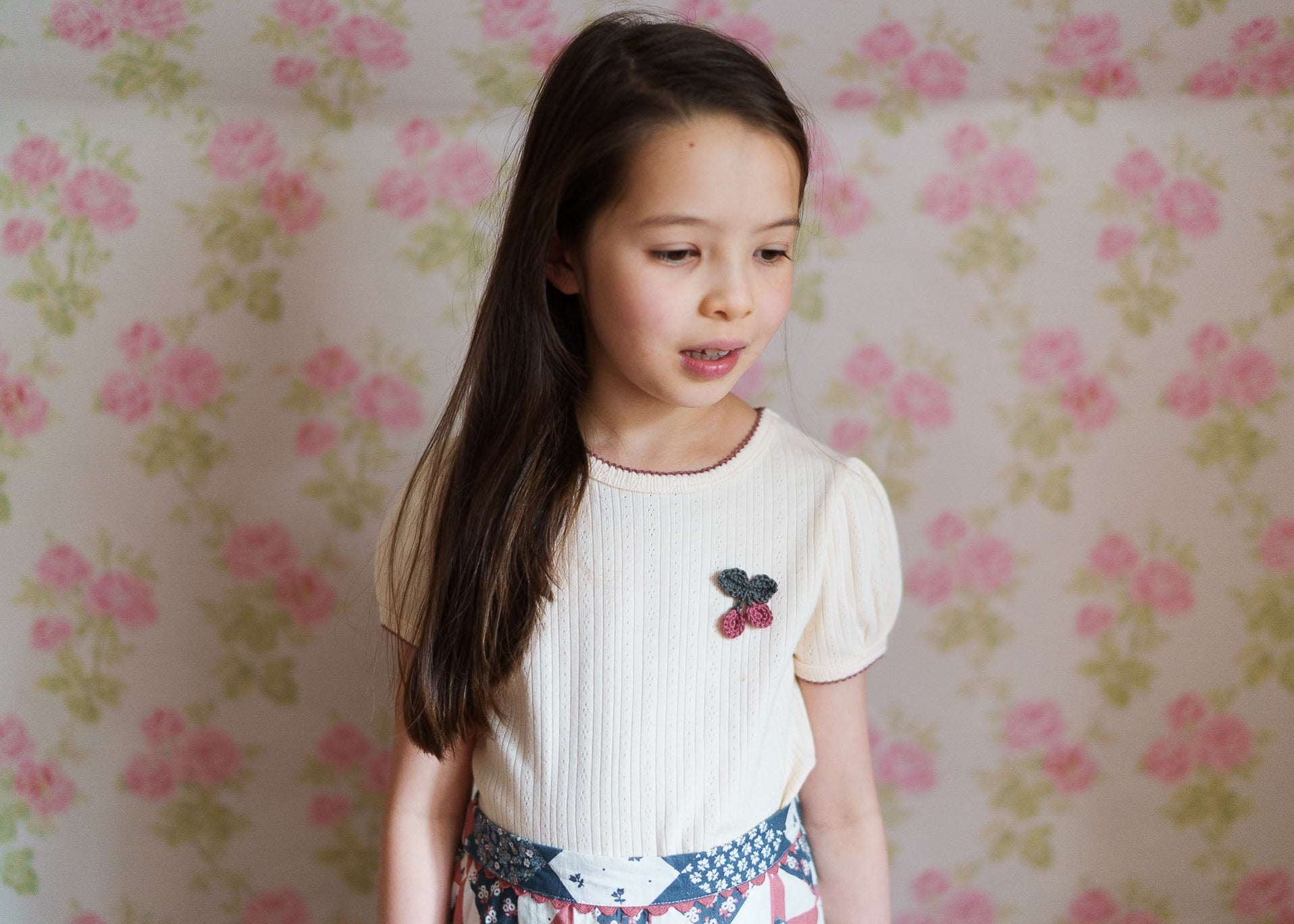 Young girl wearing a cream organic jersey cotton t-shirt with a delicate crochet cherry detail, styled in vintage-inspired children's clothing against a soft floral background.