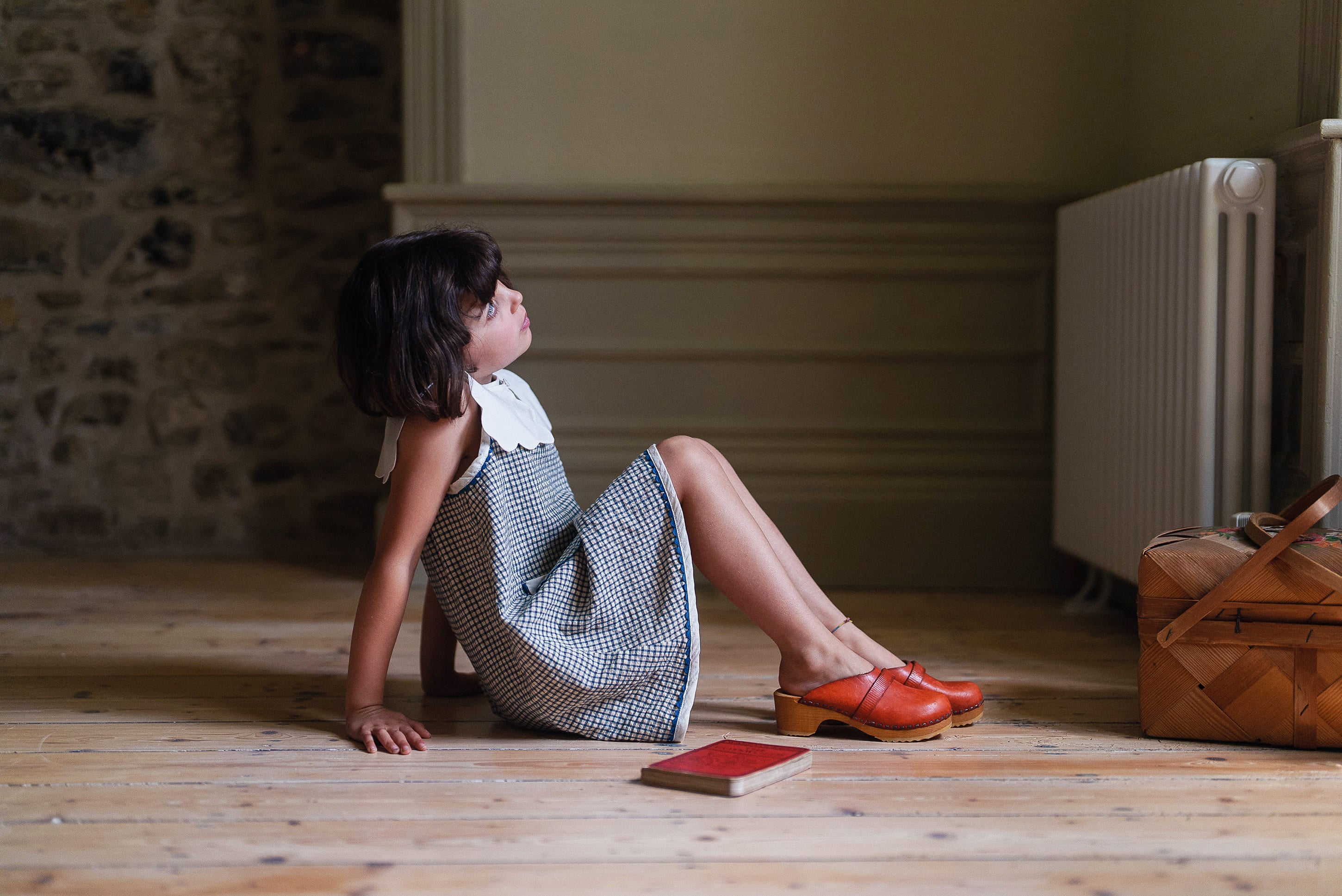 Young girl wearing a sleeveless navy and white checkered cotton dress in a vintage style, sitting on a wooden floor beside a picnic basket and a book, showcasing natural, organic children's clothing.