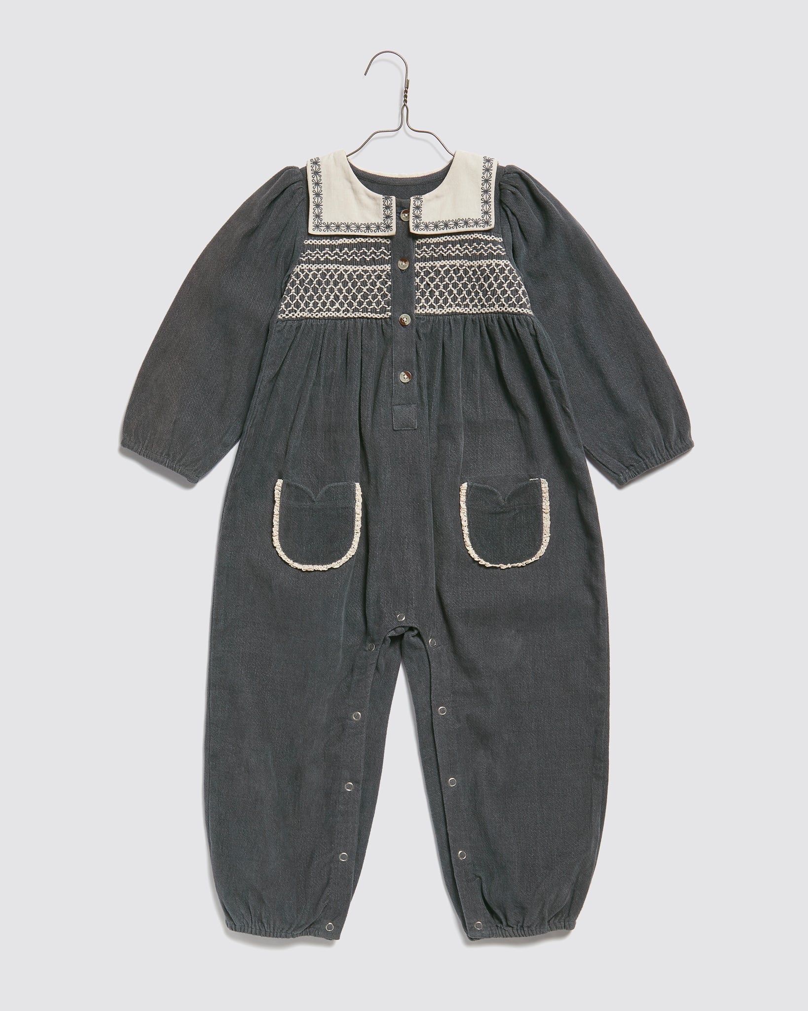 Vintage-inspired children's romper in organic cotton with long sleeves, decorative embroidery, contrast collar, and front pockets, designed for a natural, ethical look.