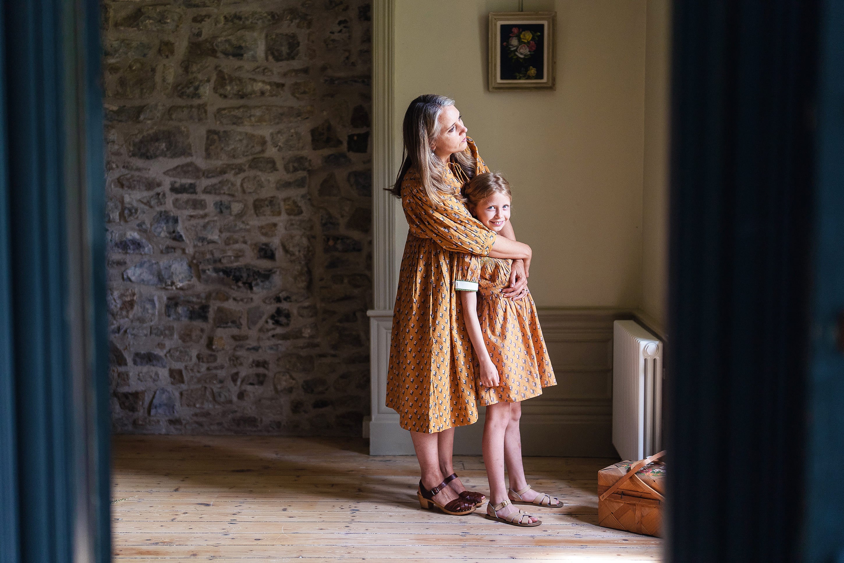Mother and daughter wearing matching saffron floral vintage-style organic cotton dresses, embracing indoors with warm natural light on wooden floors.