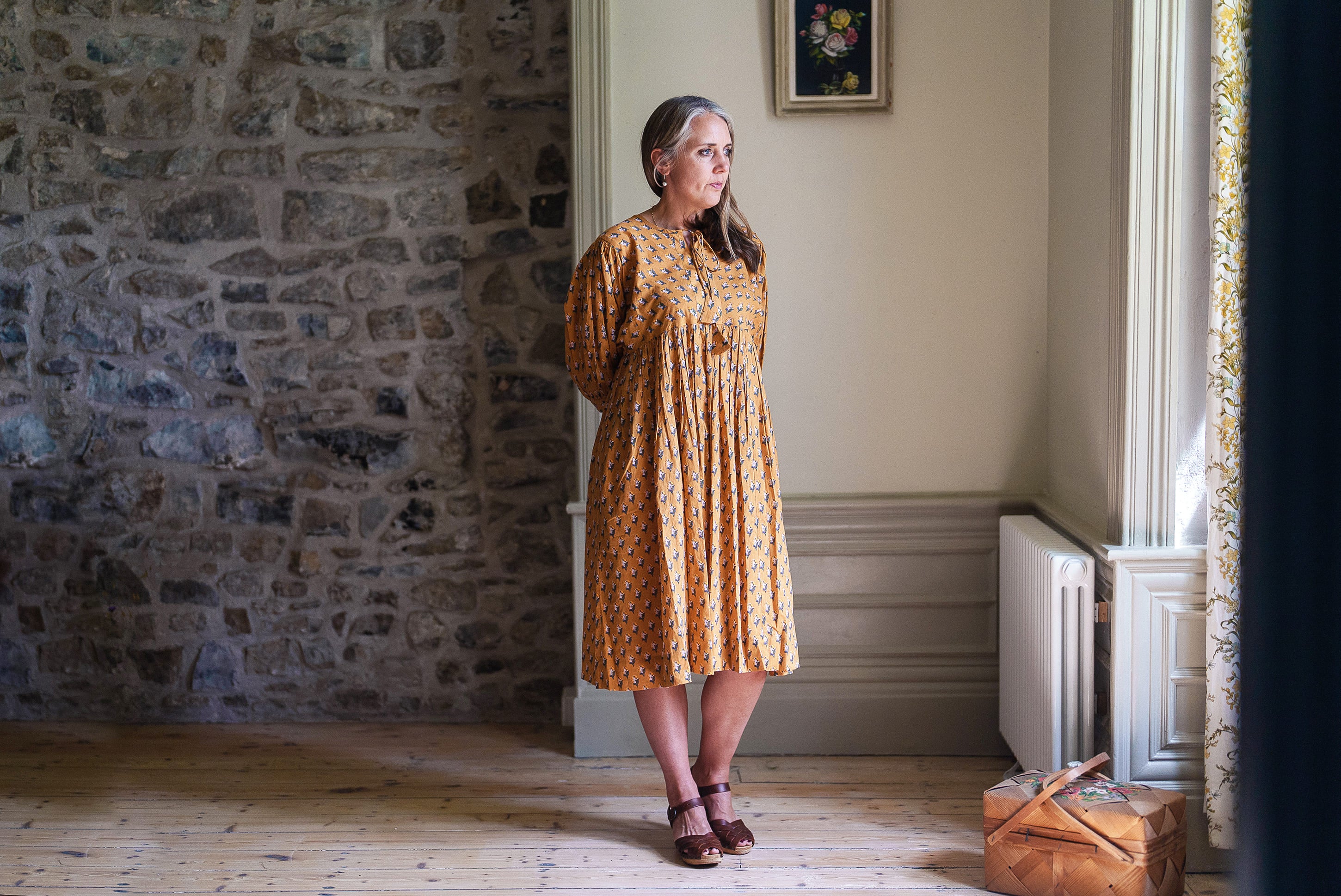 Woman wearing a vintage-style saffron floral dress made from organic cotton, standing in a bright, rustic room with natural light highlighting sustainable, ethically made clothing.