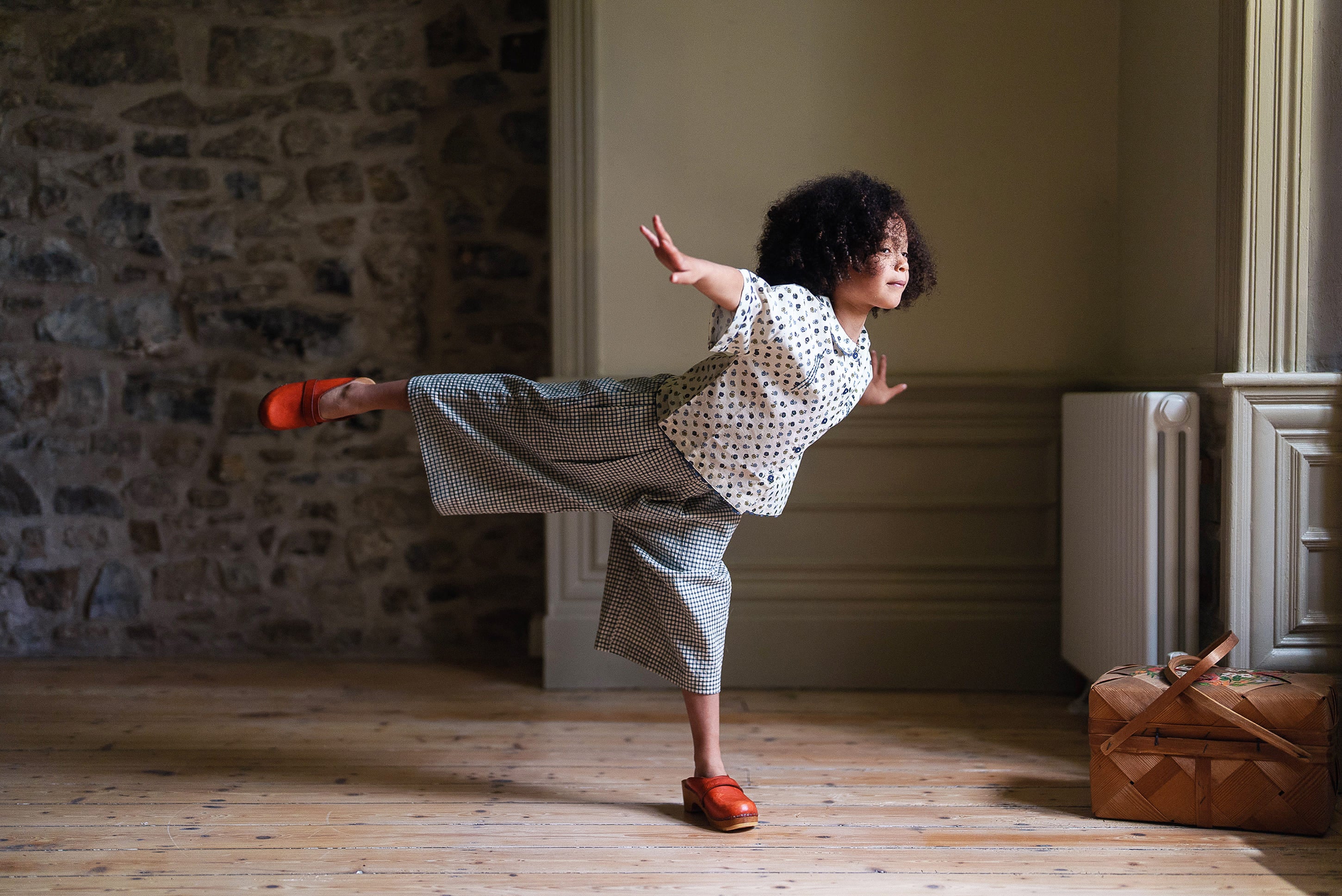 Child wearing vintage style girls cotton blouse with daisy floral print and wide pants, balancing playfully indoors in natural light near window and wooden floor.