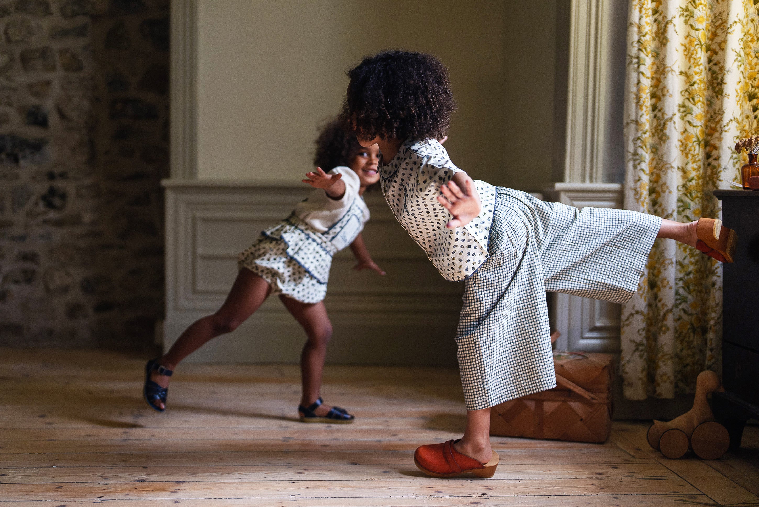 Two children playing indoors wearing vintage style, sustainable cotton clothing in a cozy, natural light-filled room with wooden floors and traditional decor.