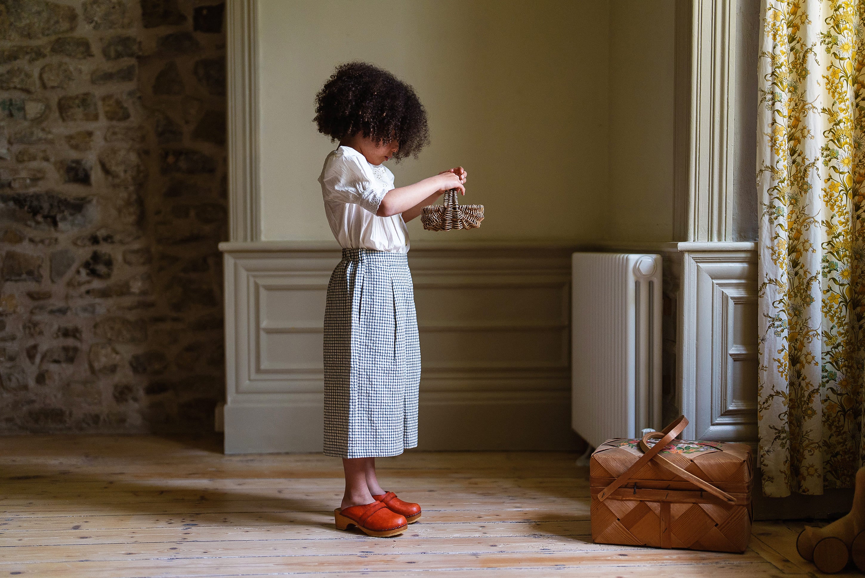 Young girl in vintage style cotton blouse and skirt standing indoors holding a small woven basket near a wooden floor and patterned curtains