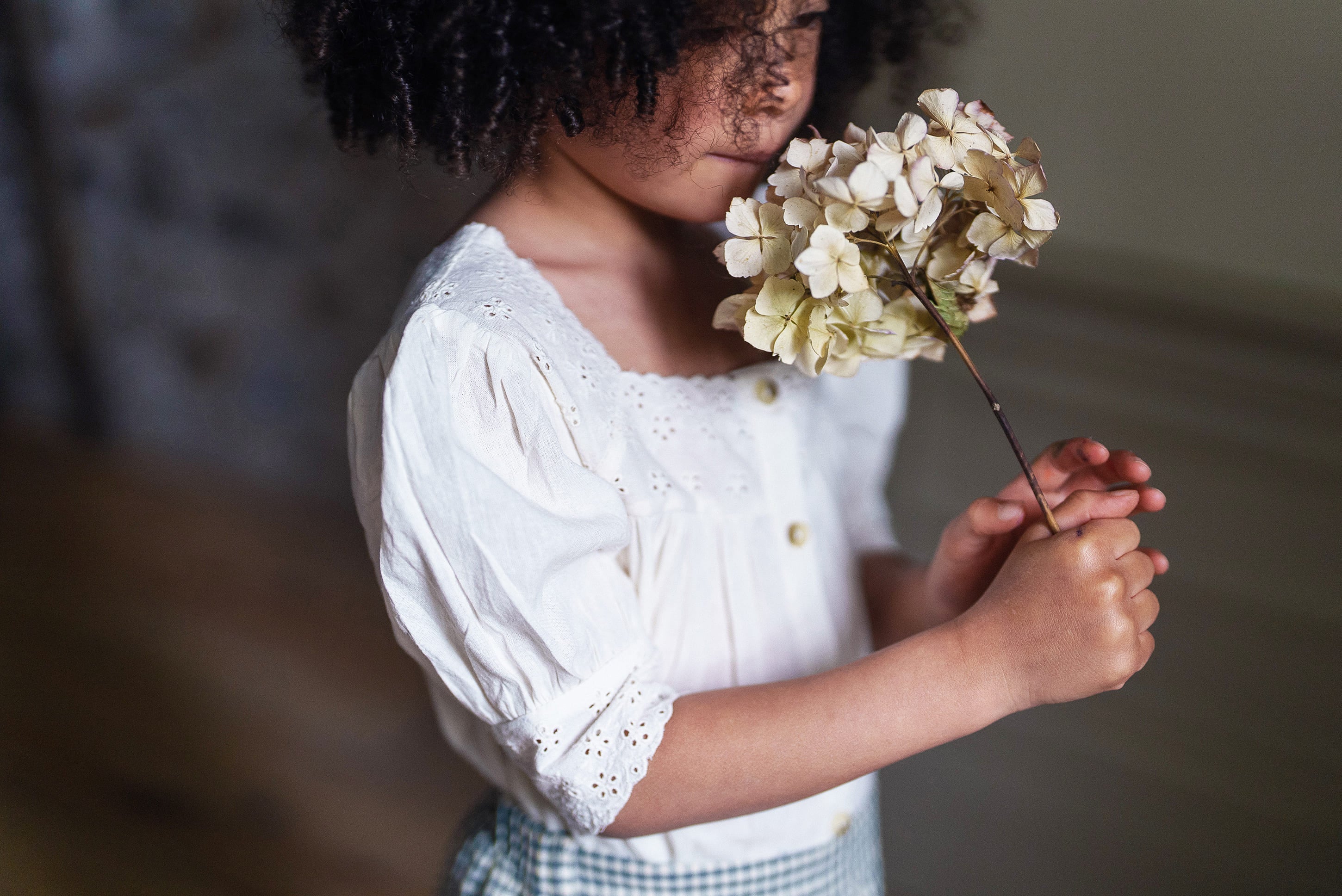 Young child wearing an off white cotton blouse with delicate embroidery, holding a dried flower, showcasing sustainable vintage style children’s clothing.