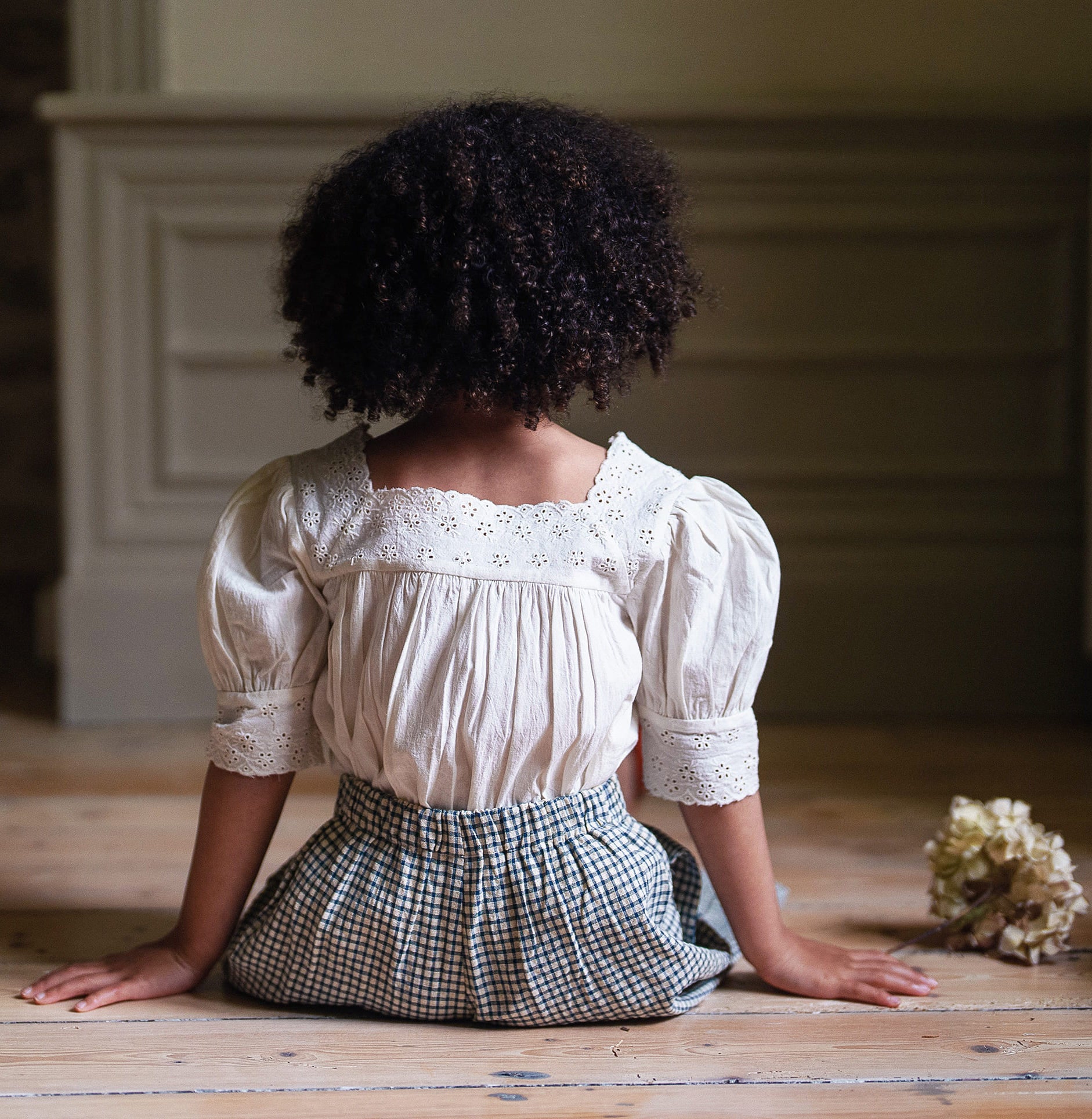 Child wearing a vintage-style off white cotton blouse with lace details and puff sleeves, paired with a checkered skirt, sitting on a wooden floor in a softly lit room.