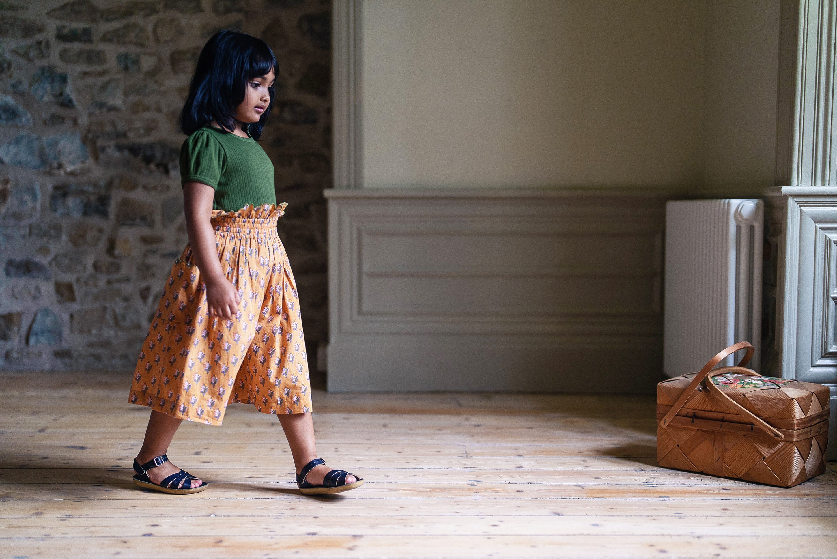 Child wearing organic cotton vintage-style green t-shirt and mustard yellow printed culottes, walking on wooden floor in softly lit traditional room with wicker basket nearby.