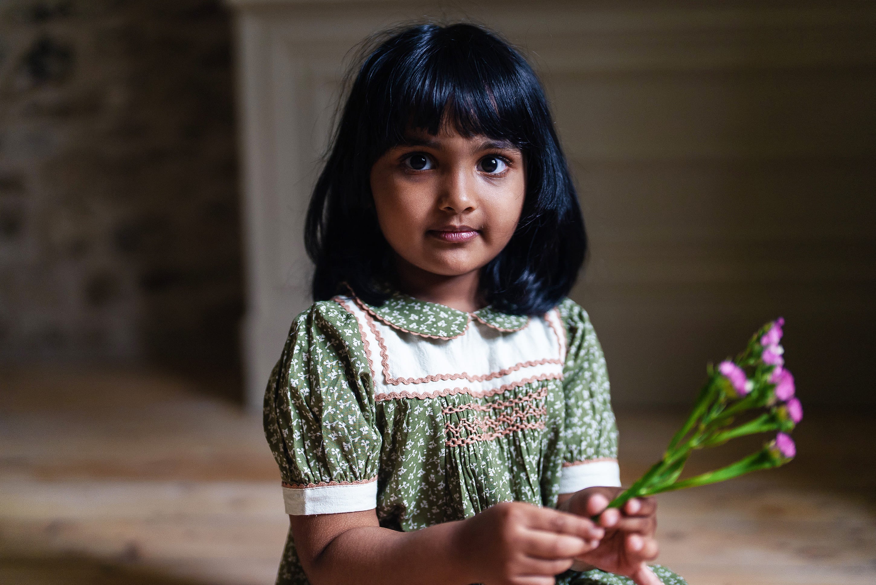 Young girl wearing a vintage style green smocked blouse with a floral pattern made from soft cotton, holding a small bouquet of pink flowers in a warm, natural setting.
