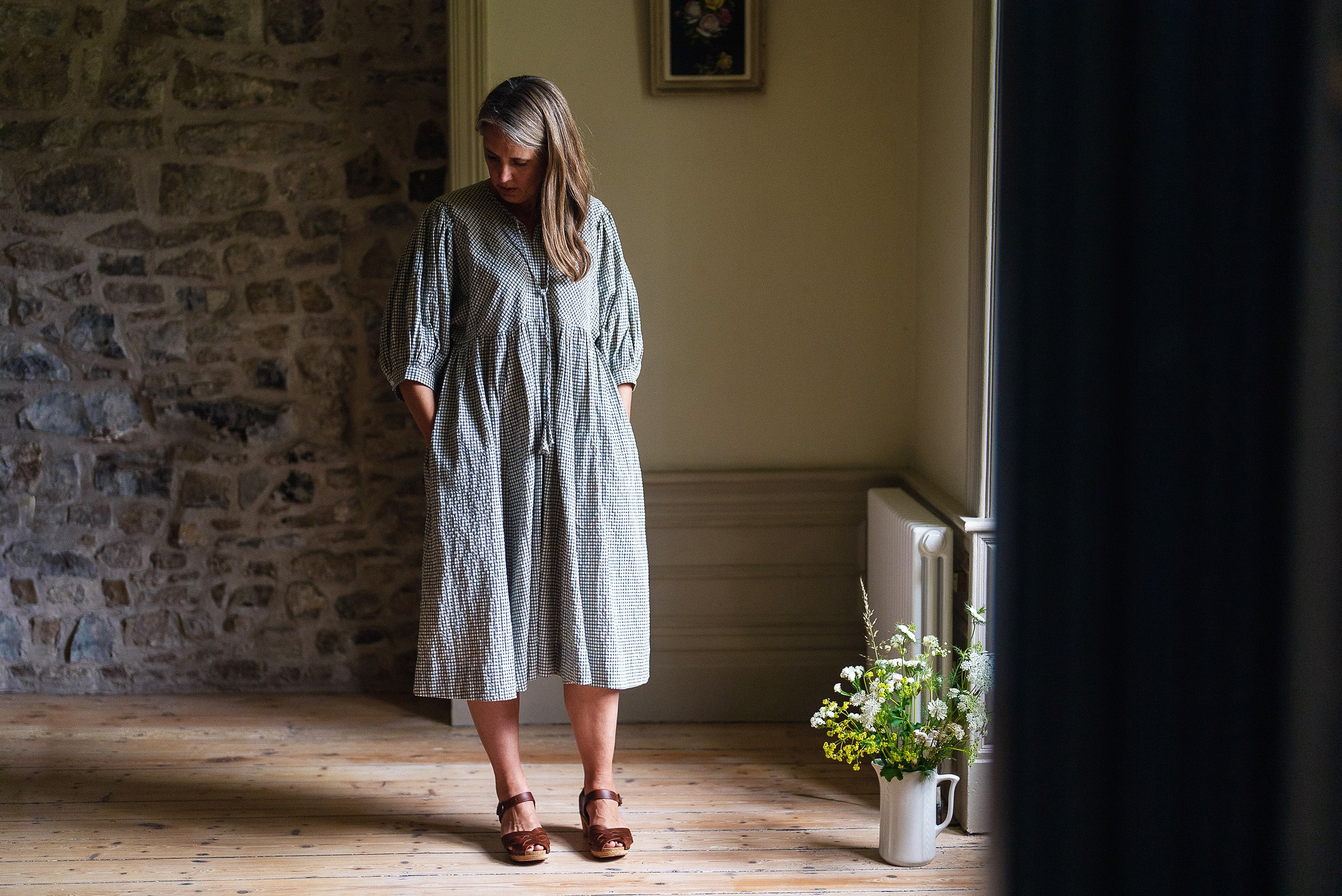 Woman wearing a natural, rustic check dress made from organic cotton standing in a softly lit vintage-style room with stone walls and wooden floors, embodying slow, ethical fashion.