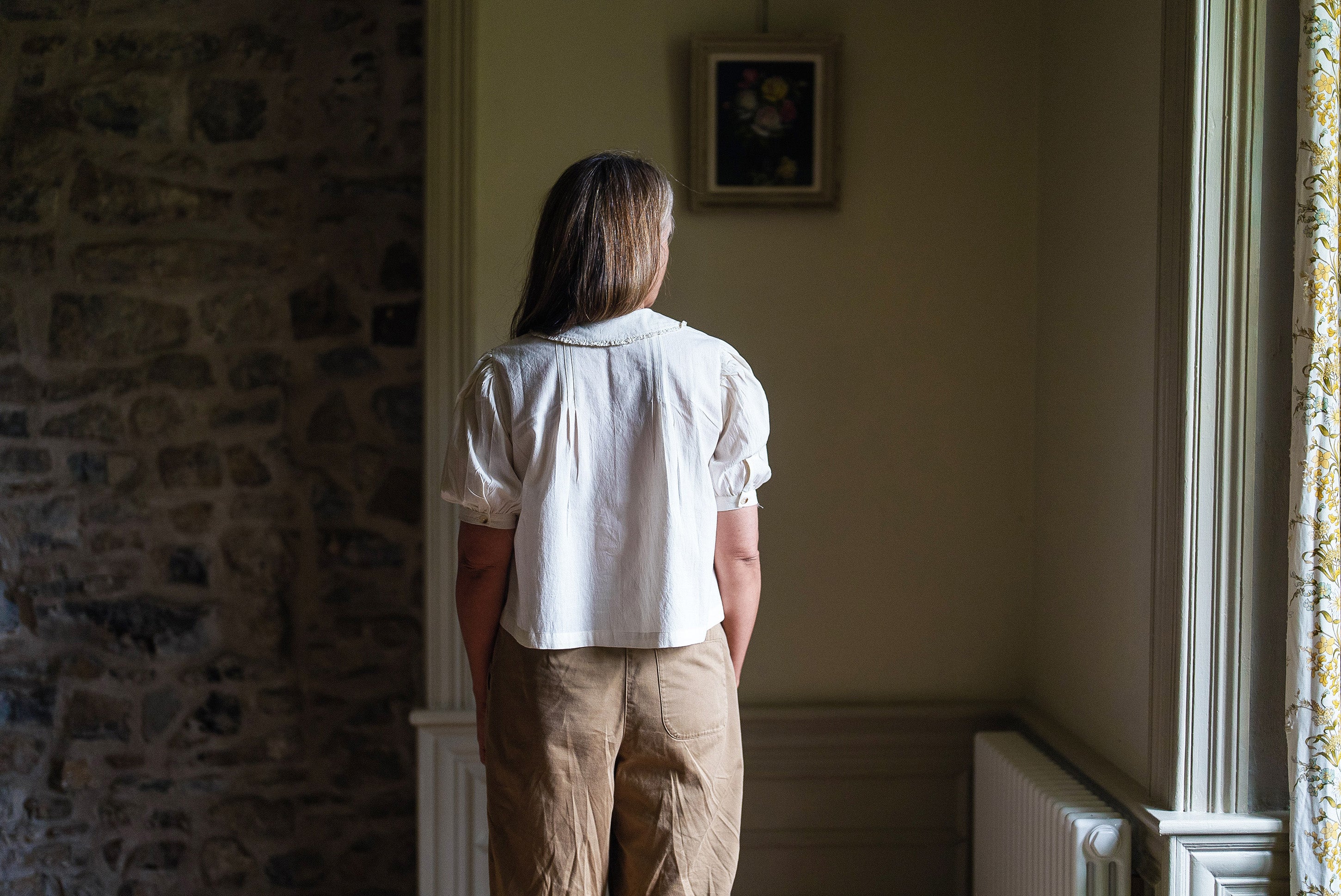 Woman standing indoors wearing a white cotton blouse with puffed short sleeves and beige pants, in a softly lit, vintage-style room with natural textures.