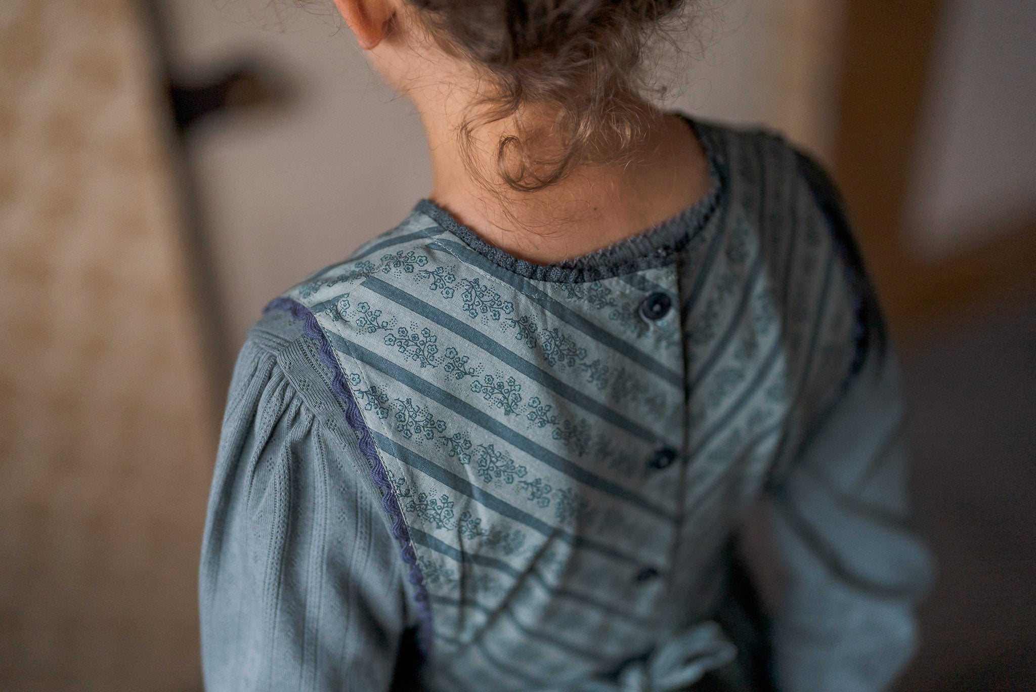 Young child wearing a vintage-style dress made from natural cotton fabric in soft slate blue tones, featuring delicate patterns and a classic buttoned back design.