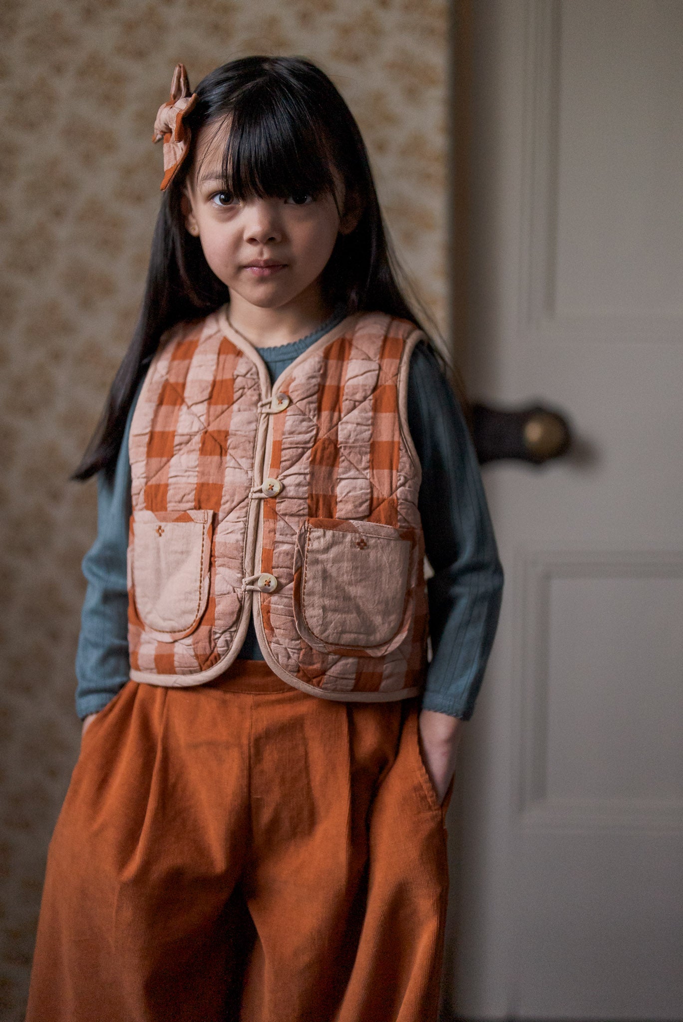 Young girl wearing a navy blue organic cotton top with a vintage-style orange checkered quilted vest and loose orange pants, showcasing sustainable, ethically made children’s clothing.