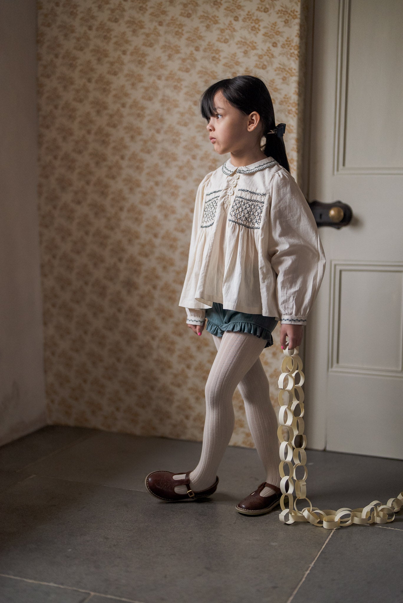 Young girl wearing vintage style organic cotton blouse and slate blue bloomers with natural ribbed tights and brown leather shoes in a softly lit room with floral wallpaper.
