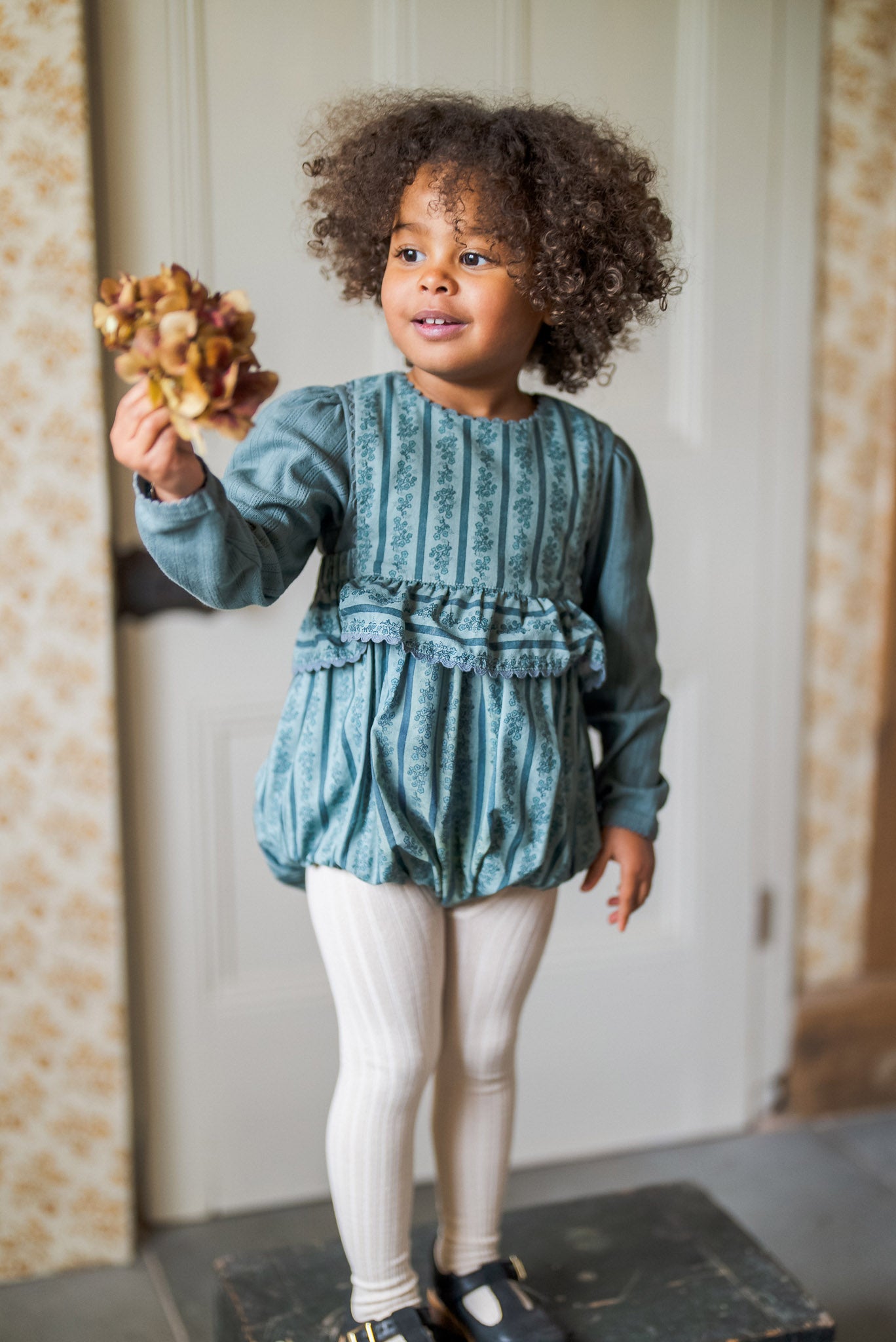 Young child wearing vintage style cotton dress with traditional floral pattern and white ribbed tights inside a cozy home, embracing slow, natural, ethical children’s clothing.