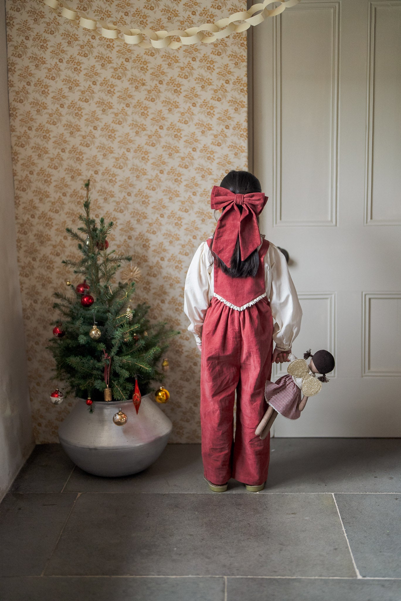Child wearing vintage style red velvet bow clip and matching outfit, holding a doll, standing by a small decorated Christmas tree in a cozy, softly lit room.
