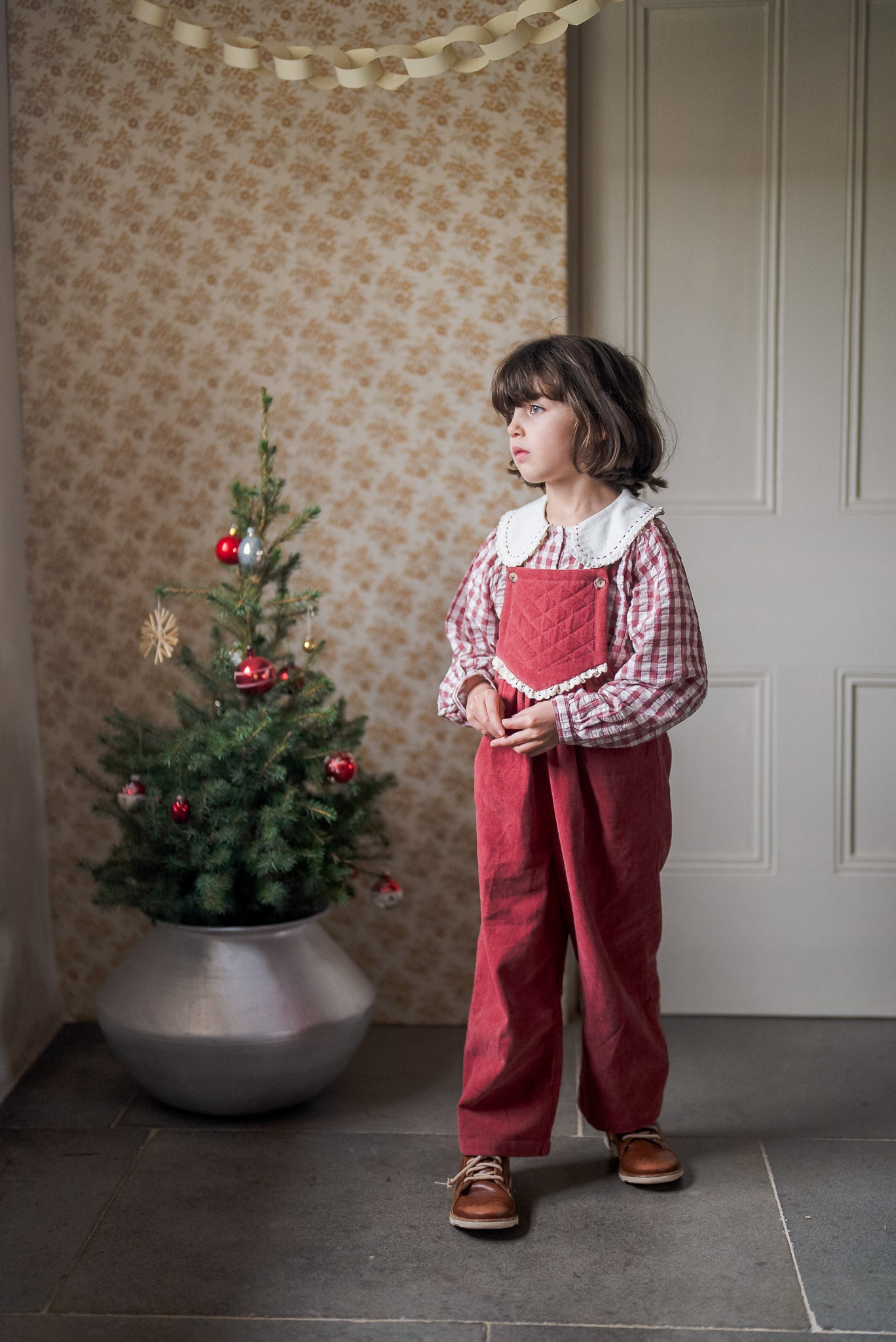 Young girl wearing a vintage-style red and white checkered cotton blouse with red overalls, standing indoors beside a small decorated Christmas tree in a natural, softly lit room.