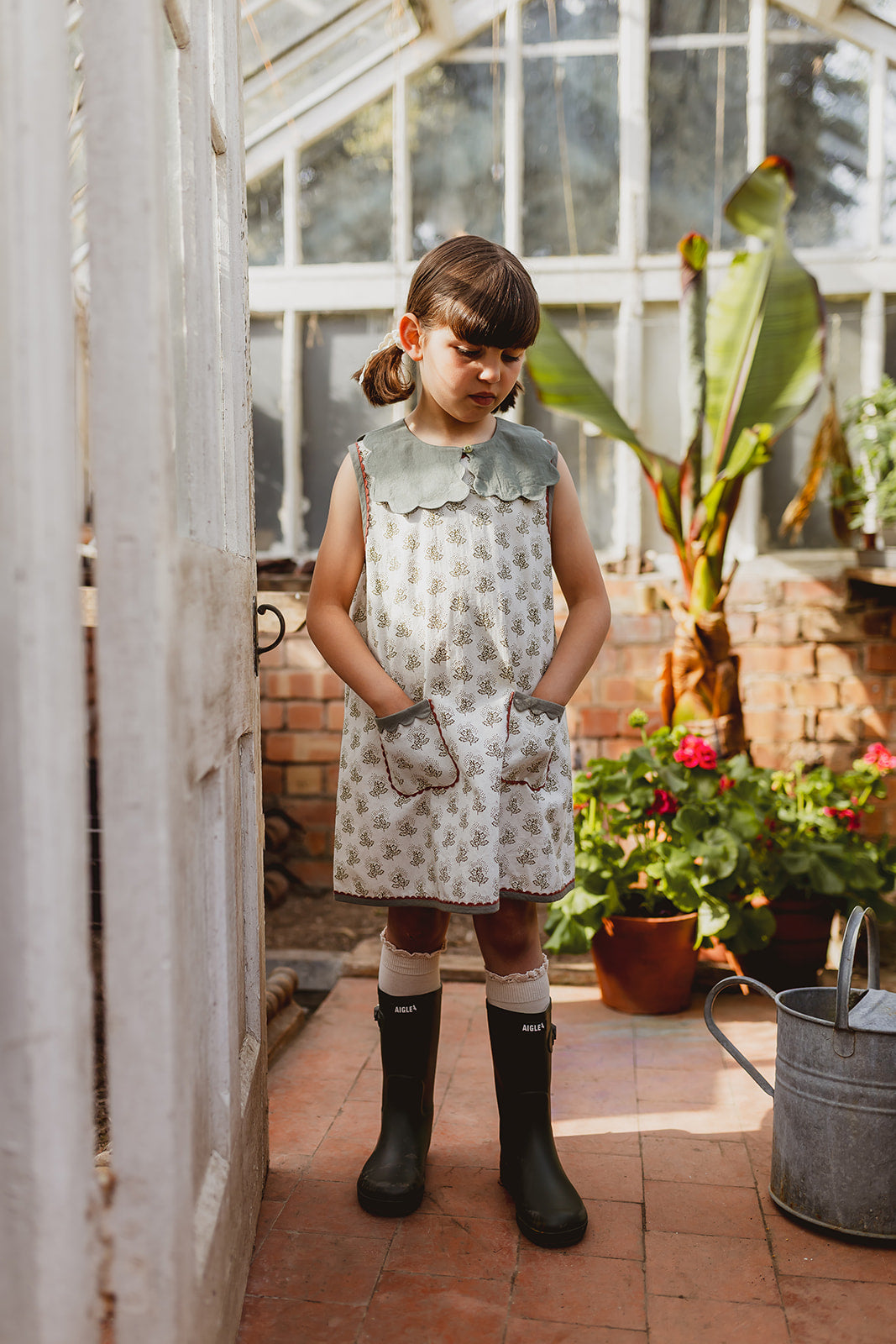 Young girl in a vintage style organic cotton dress with a scalloped green collar standing in a sunlit greenhouse surrounded by plants and gardening tools.