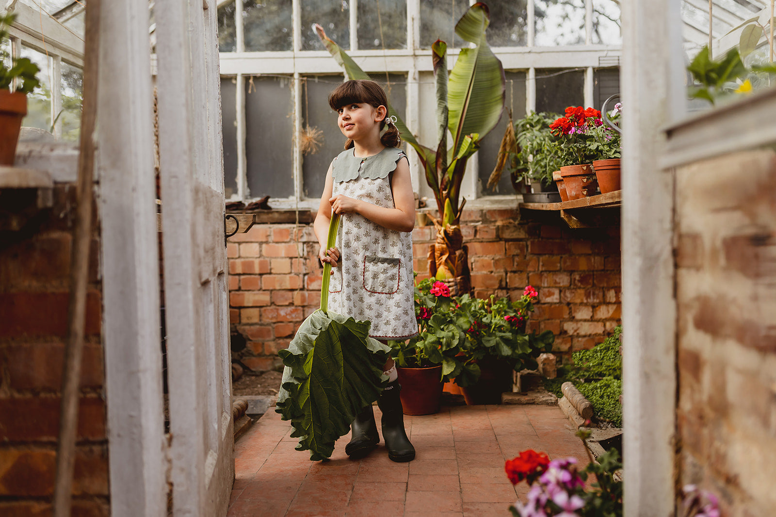 Young girl wearing a vintage-style organic cotton dress with a scalloped collar in green, standing in a greenhouse surrounded by plants and flowers, showcasing sustainable children's clothing.