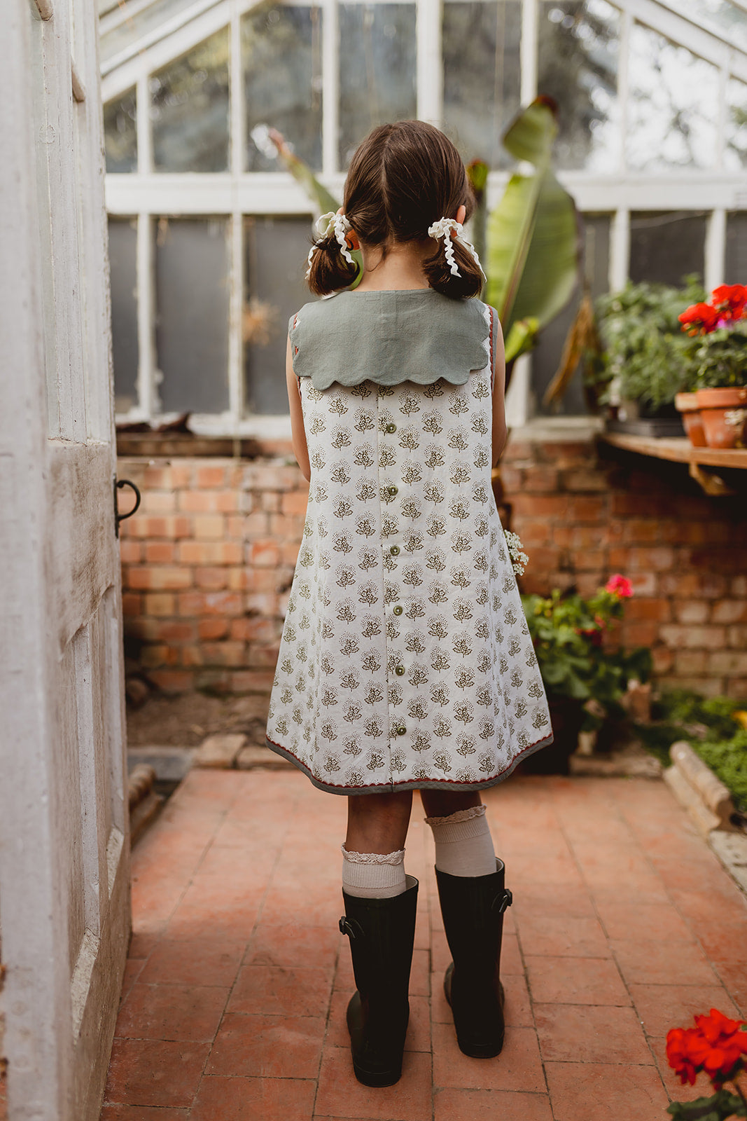Young girl wearing a vintage style organic cotton dress with a green scalloped collar, standing in a garden room, showcasing sustainable and ethically made children's clothing.