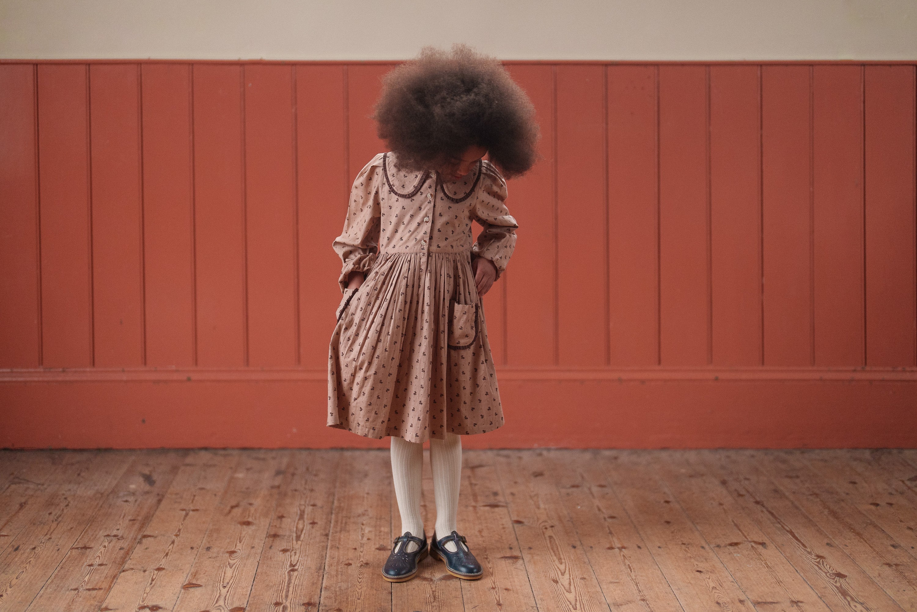 Child wearing a vintage style pink floral organic cotton dress with long sleeves and a Peter Pan collar, standing on a wooden floor against a muted terracotta wall.