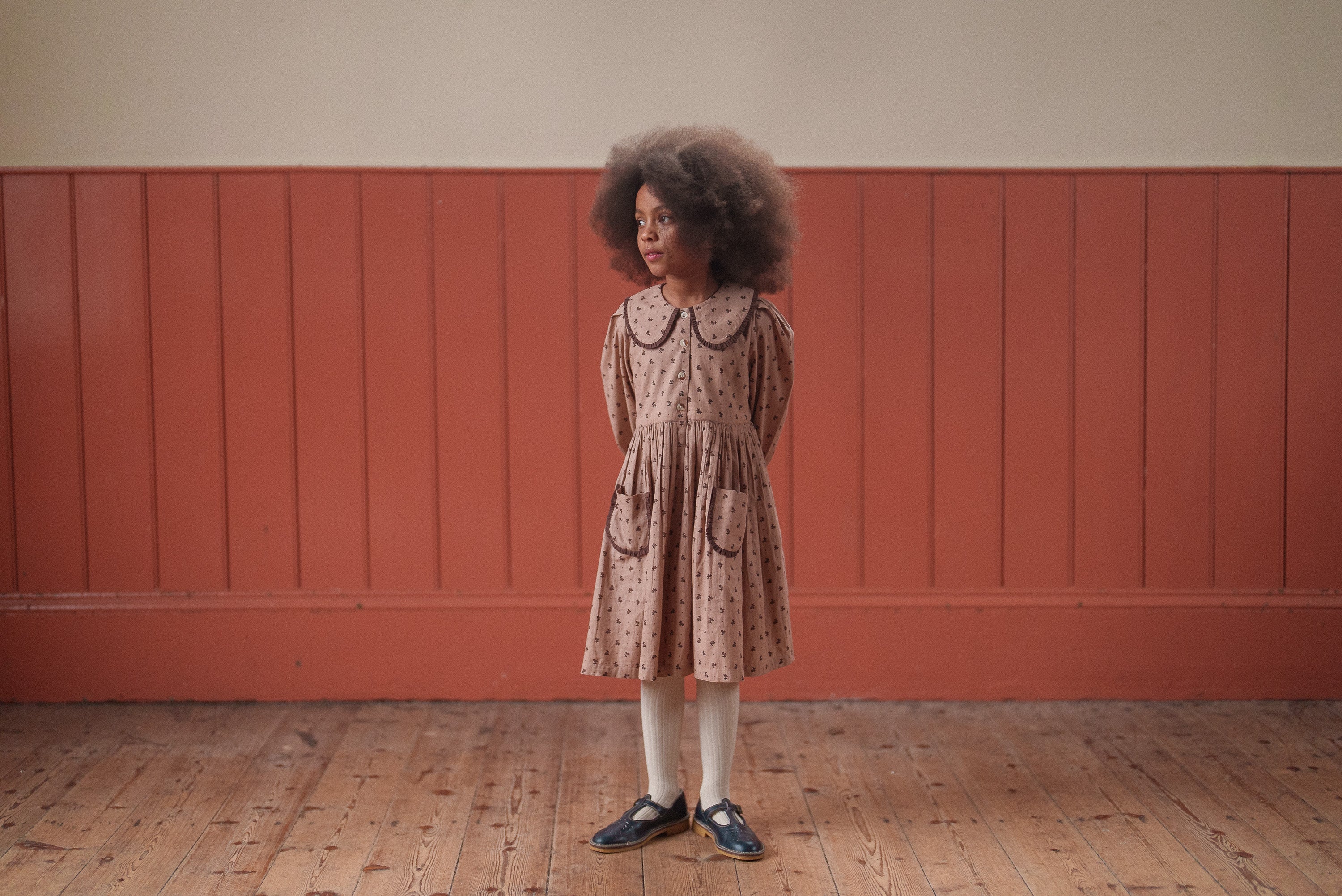 Young girl wearing a pink and brown vintage-style organic cotton dress with floral pattern, paired with white tights and black shoes, standing in a warm, rustic room.