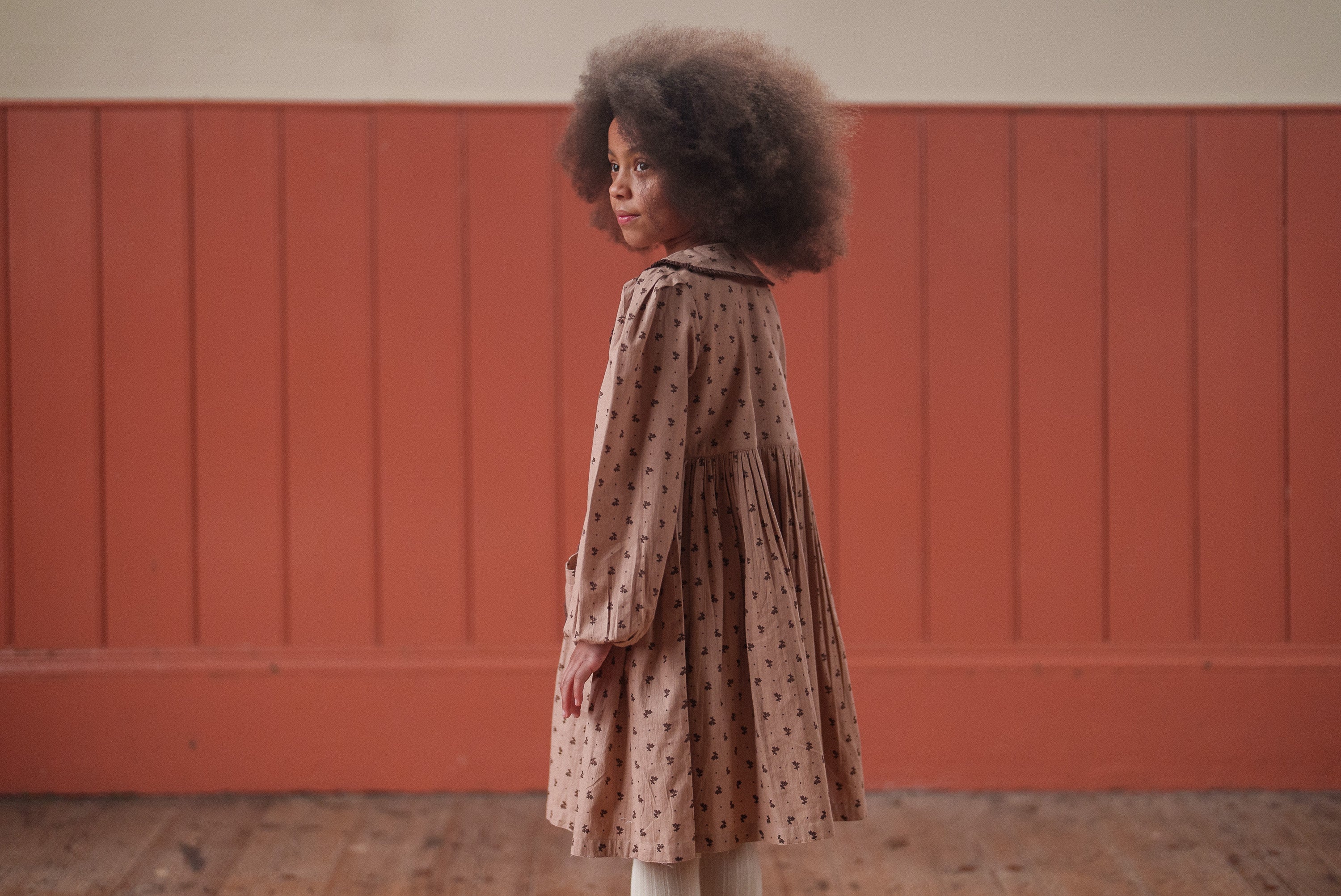 Young girl wearing a vintage-style dress in soft pink with brown floral patterns, made from organic cotton, standing against a warm-toned wall in a natural light setting.