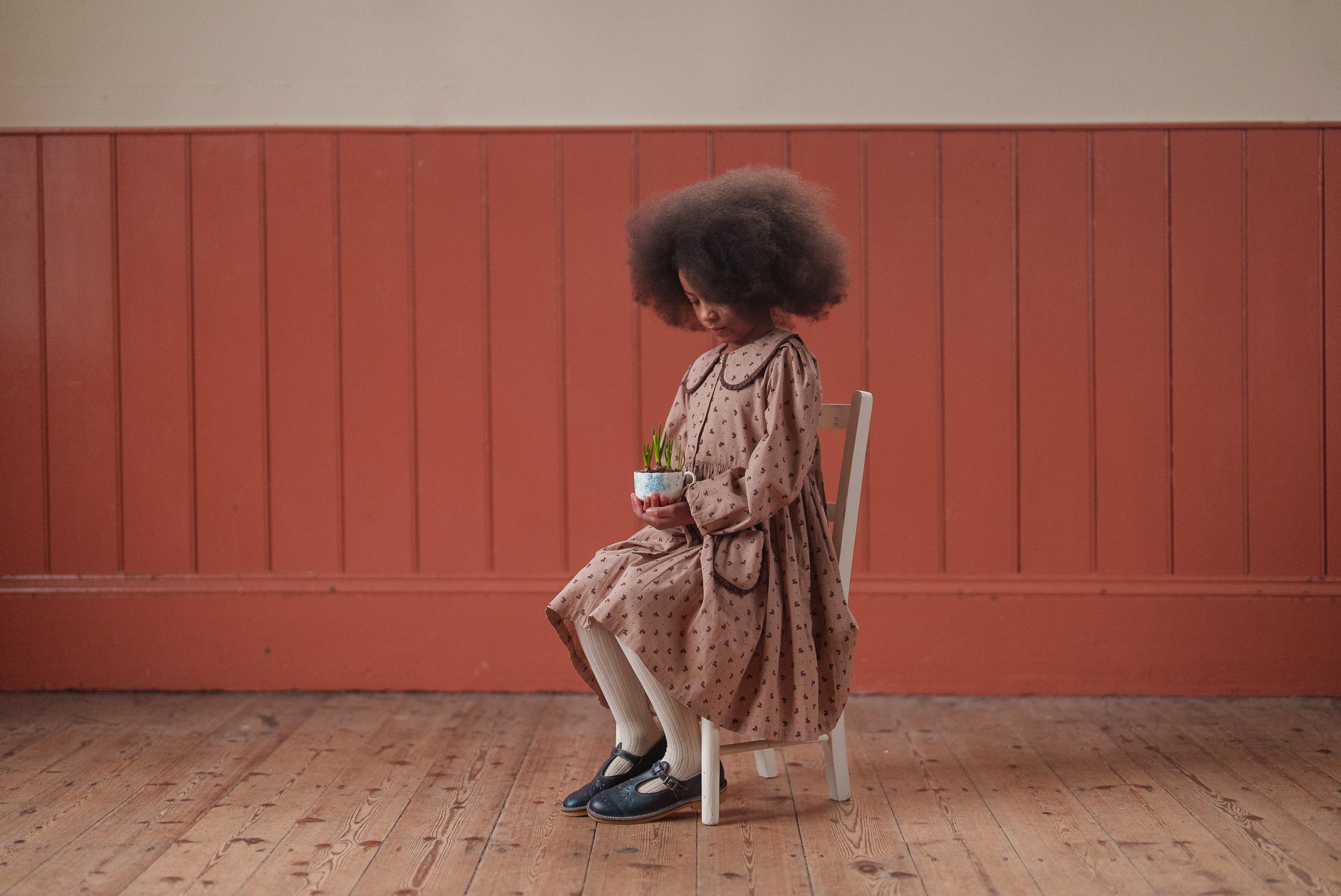 Young girl wearing a vintage style pink floral organic cotton dress sitting on a white wooden chair holding a small plant, in a warm, natural indoor setting.