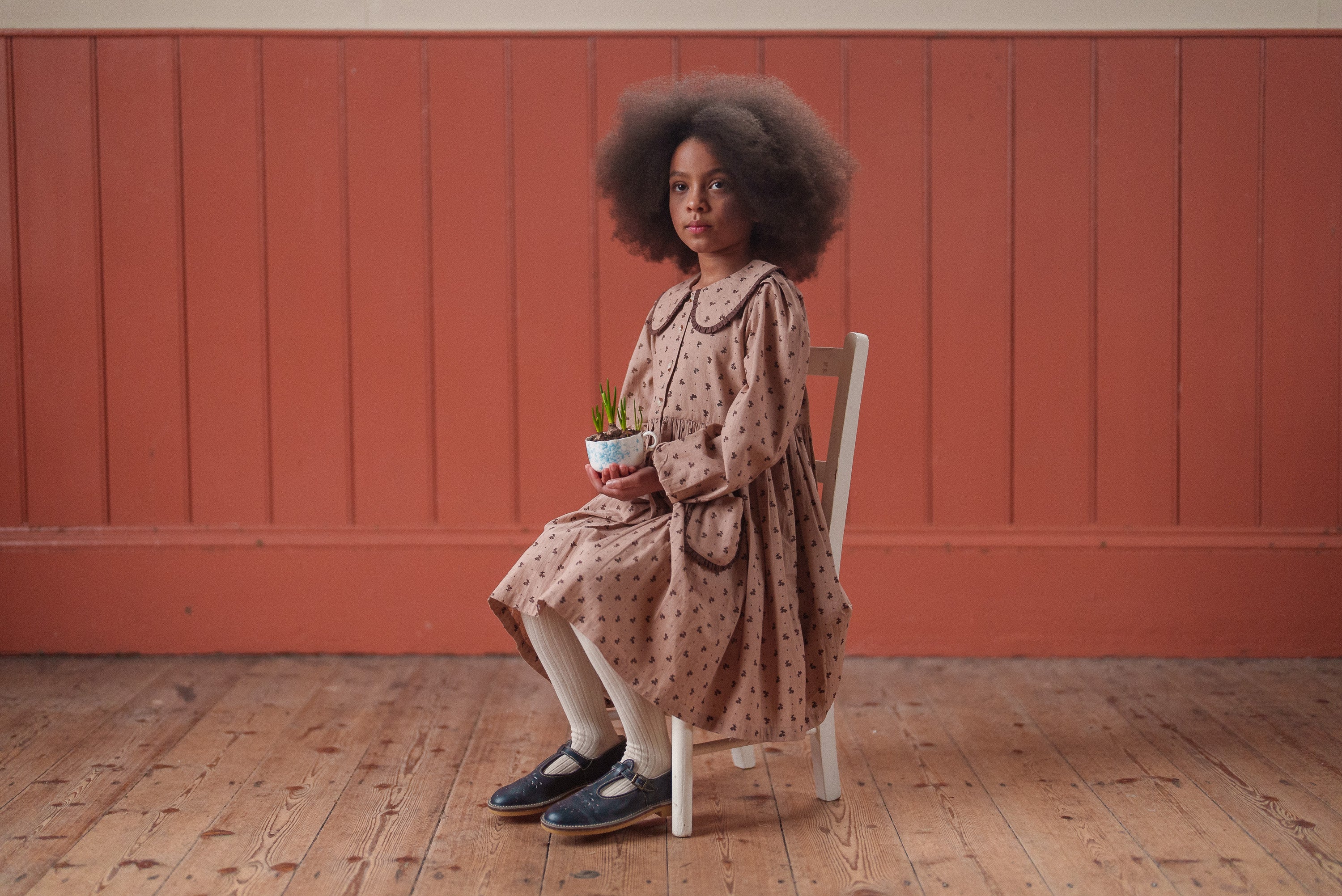 Young girl wearing a vintage style dress in organic cotton with a floral pattern sitting on a wooden chair holding a small plant, showcasing sustainable children's clothing.