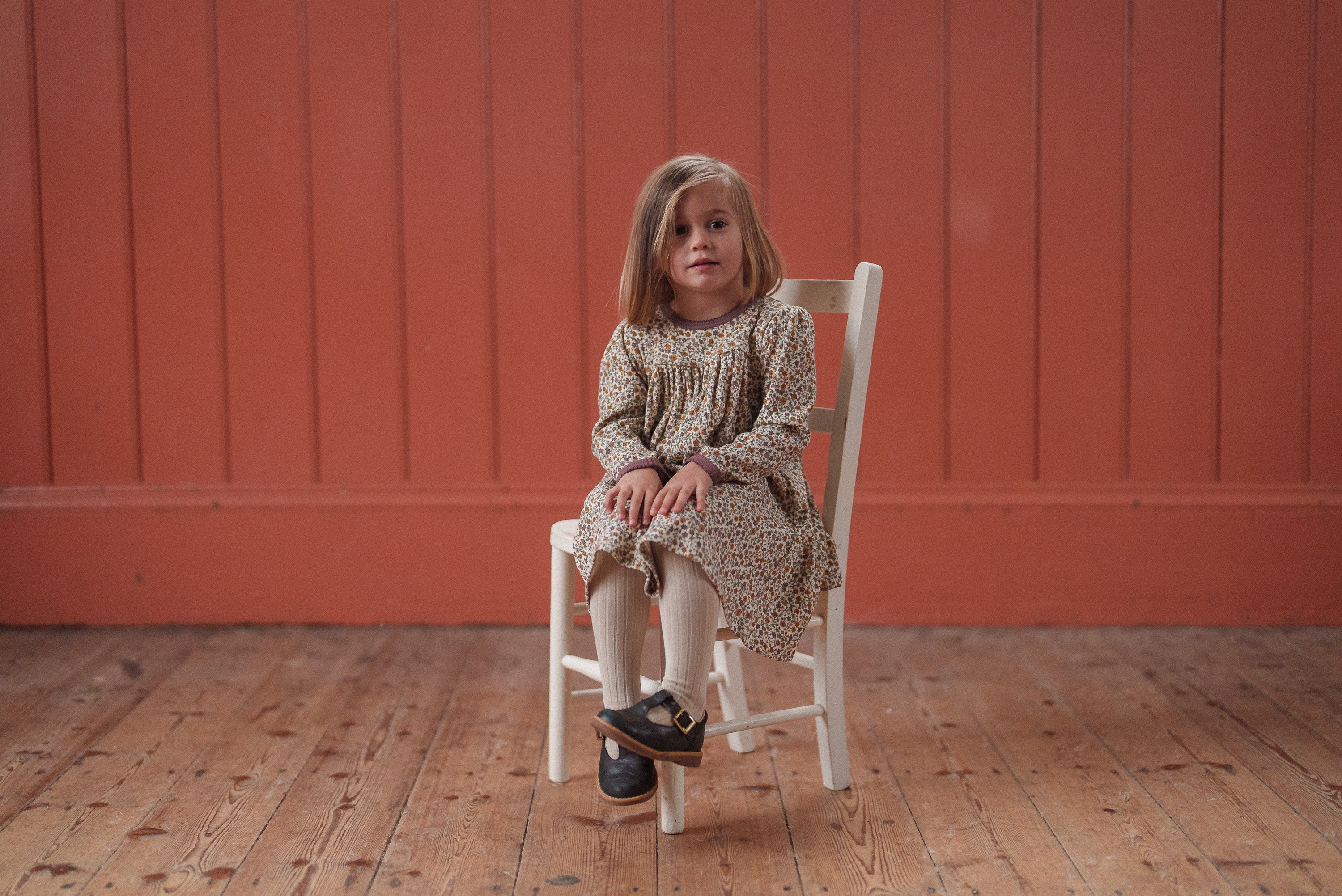 Young girl wearing a vintage style organic cotton floral dress sitting on a white wooden chair against a natural wood floor and warm orange wall background.