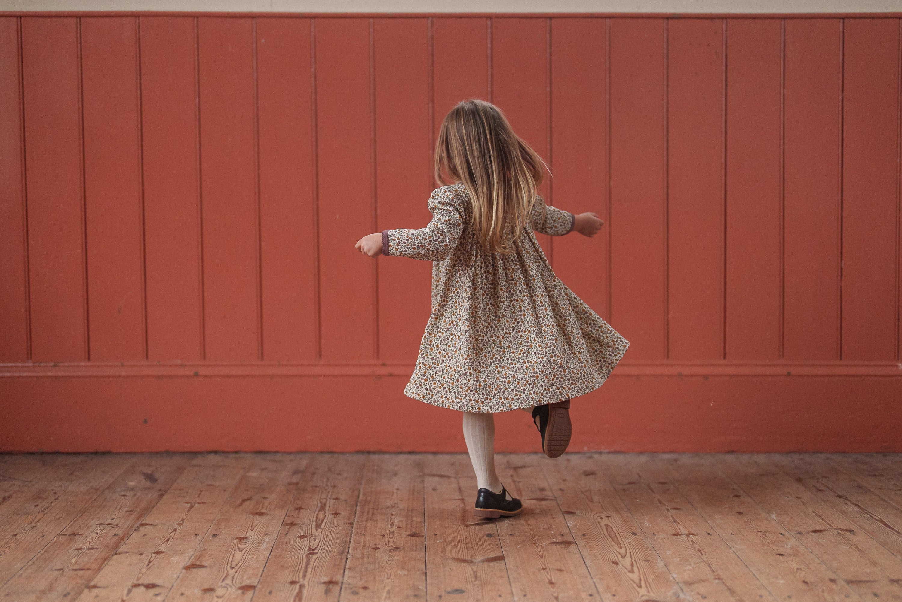 Young girl twirling in a vintage-style, organic cotton dress with a multicolor prairie floral pattern, set against a warm, rustic wooden floor and red wall.