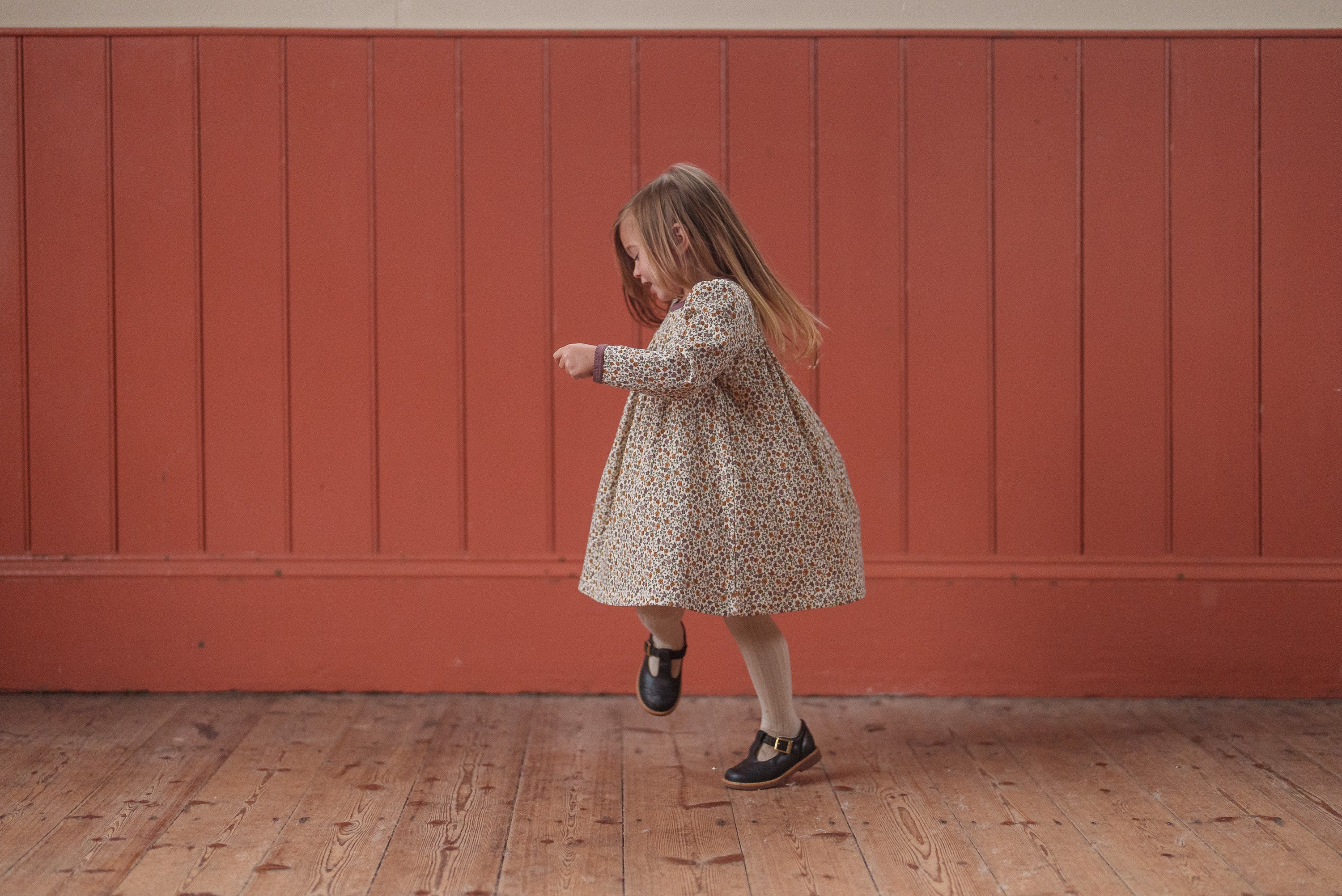 Young girl wearing a vintage-style organic cotton dress with a subtle floral pattern, playing indoors on a wooden floor against a warm-toned wall.