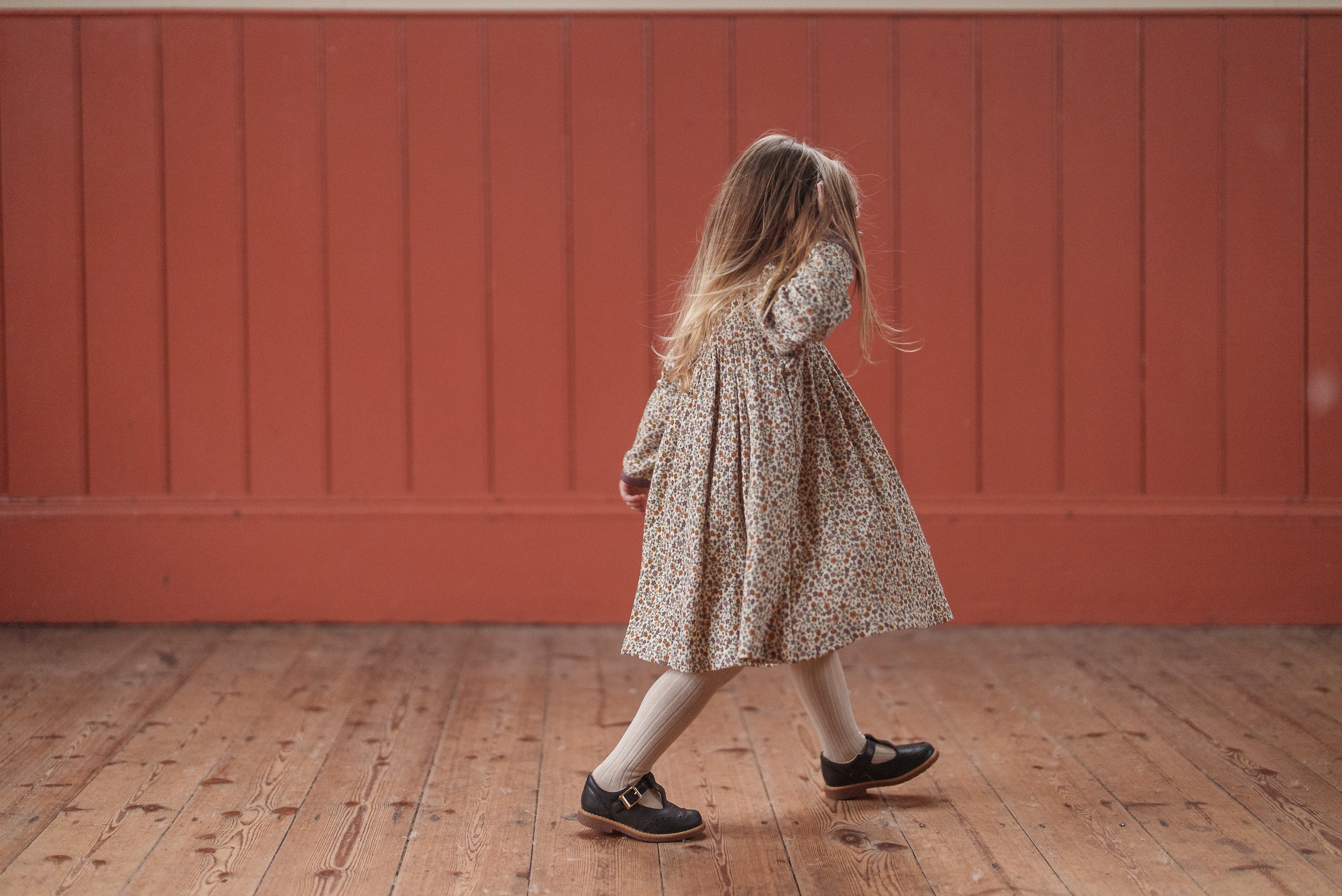 A young girl wearing a vintage style, organic cotton dress with a multicolor floral pattern, walking on a wooden floor in a warm, natural setting.