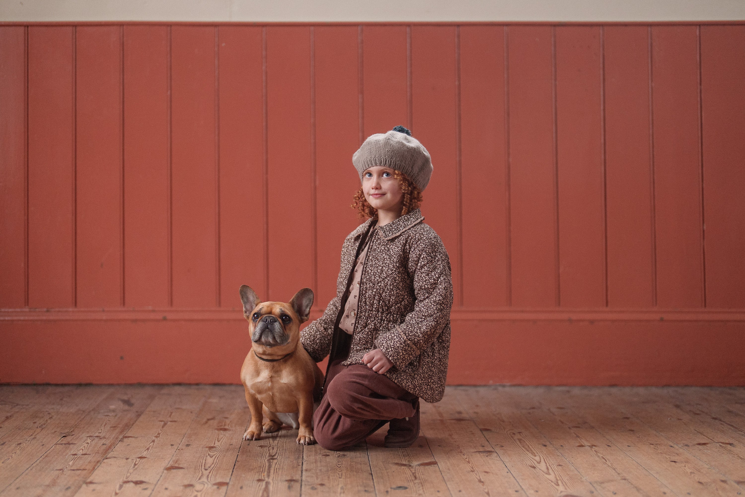 Young child wearing a vintage-style outfit with a hand-knitted merino beret in oat color, posing indoors with a small dog, showcasing natural, sustainable kids clothing.