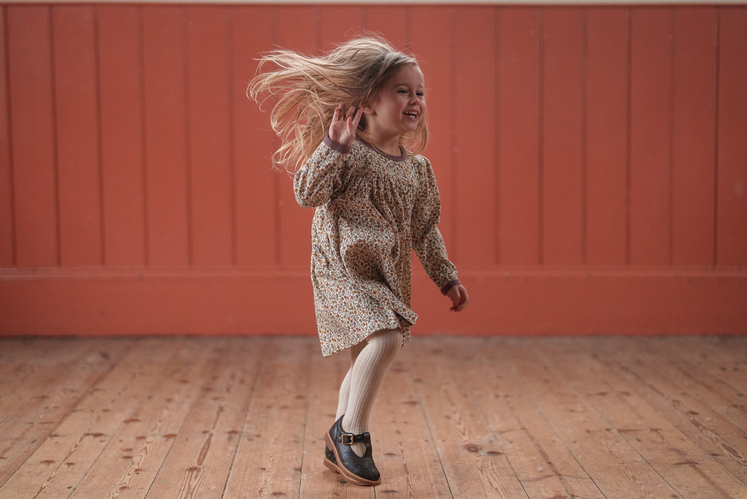 A young girl joyfully twirling in a vintage-inspired organic cotton dress with prairie floral print, paired with tights and classic shoes, in a warm, natural setting.