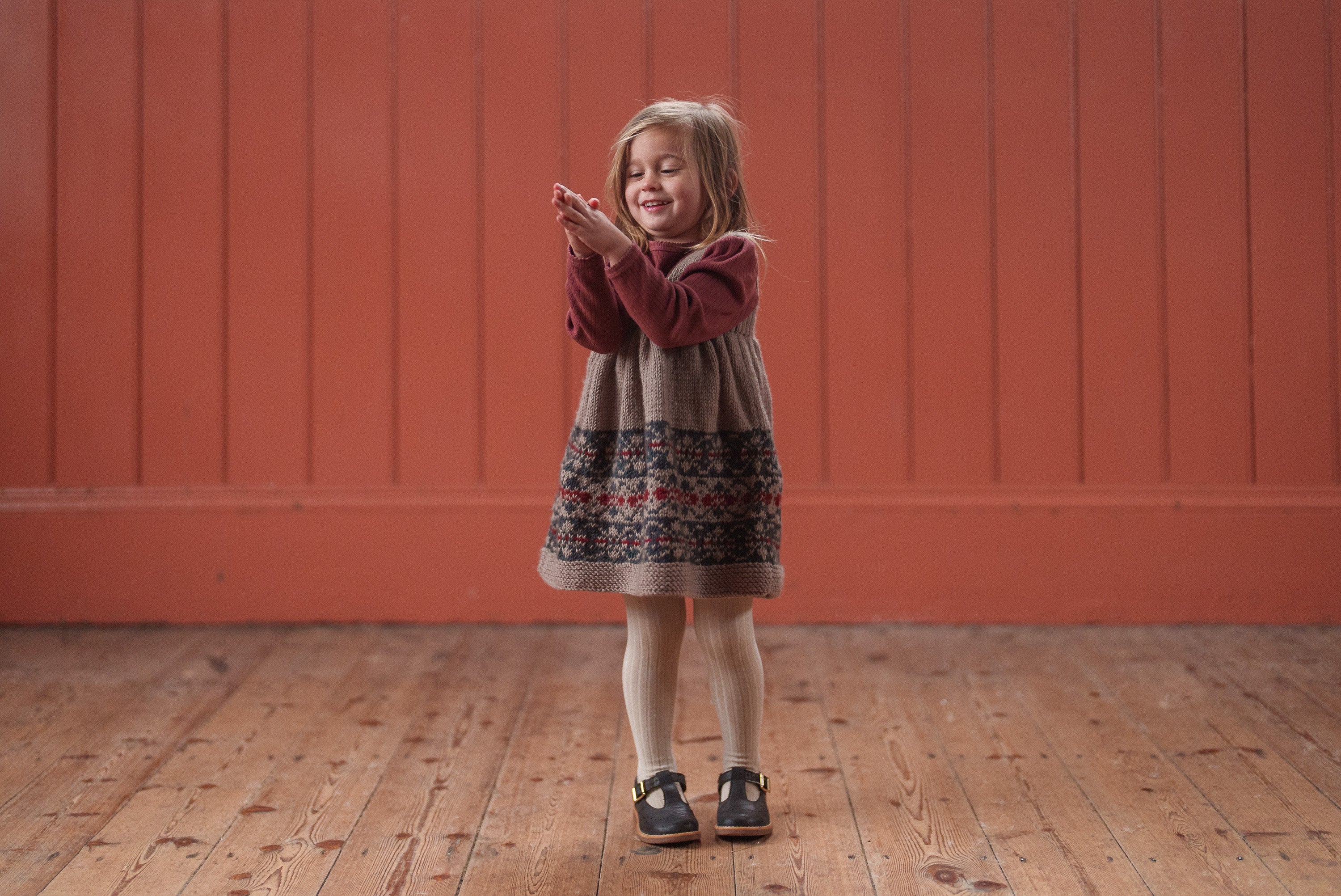 A young girl wearing a hand-knitted pinafore dress with Fair Isle pattern in navy, blue, beige, and multicolor, standing on a wooden floor with a warm smile in a vintage-style setting.