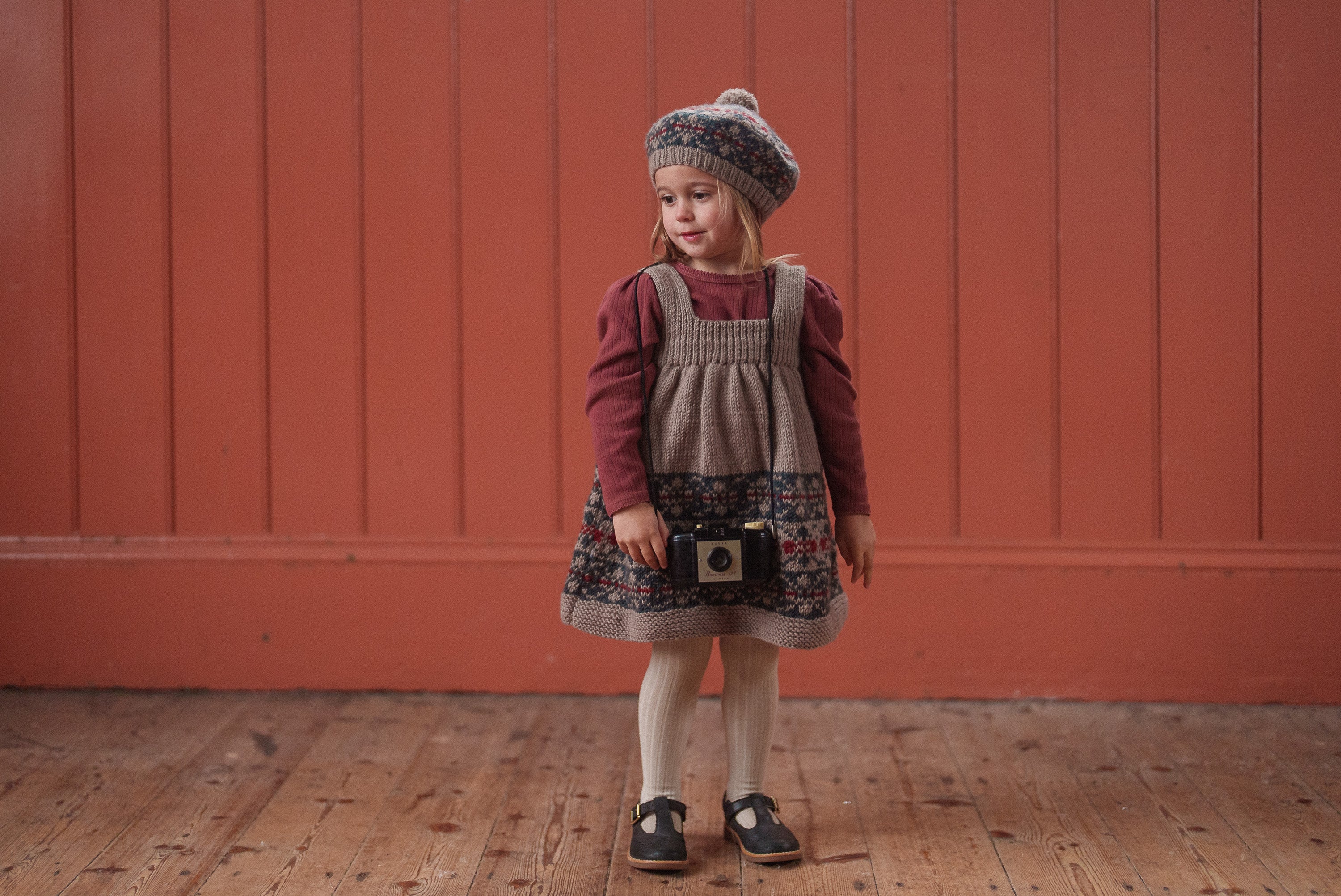 A young girl wearing vintage style sustainable children's clothing with a rich red organic cotton top and a patterned pinafore dress, standing against a warm red wooden wall.