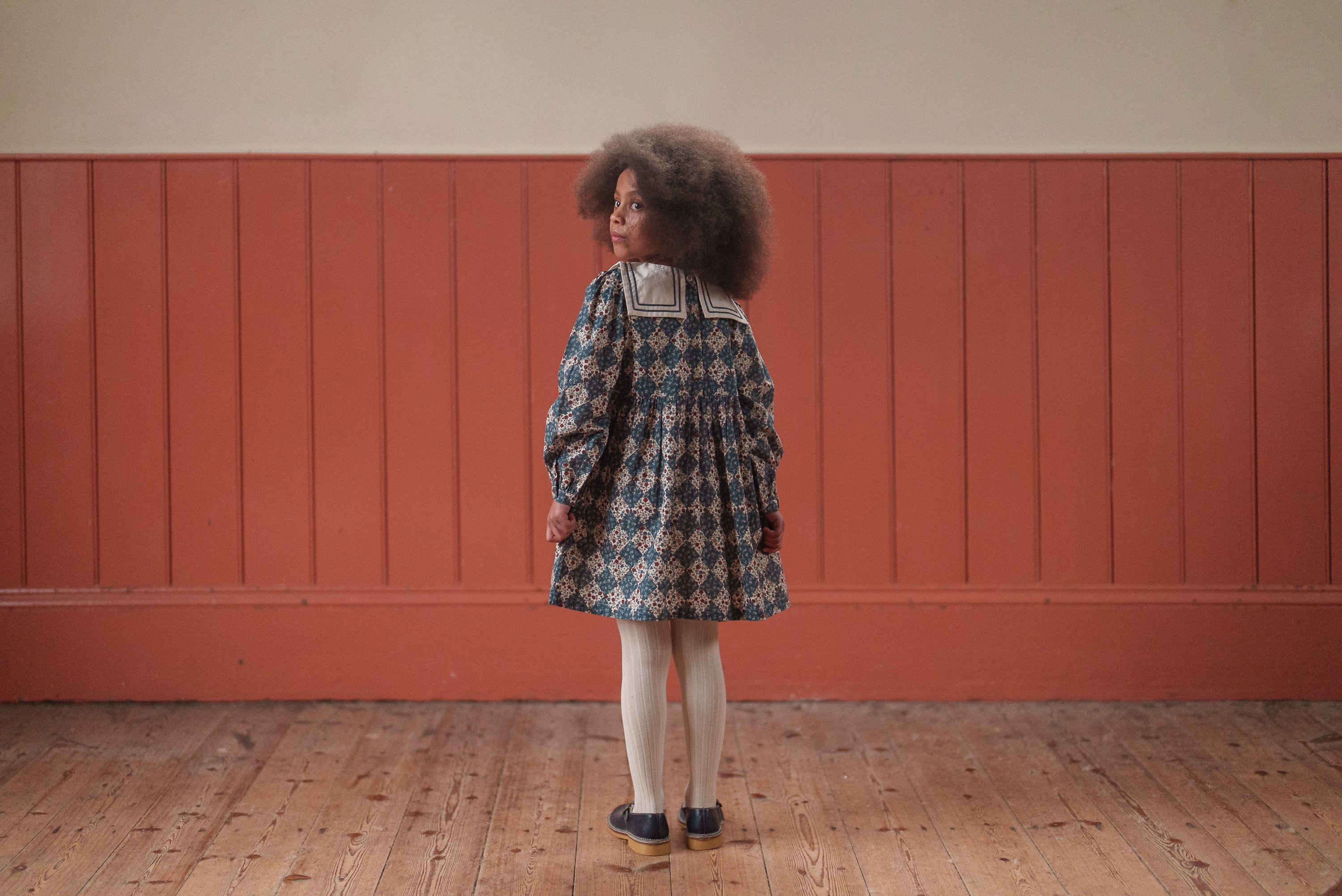 Young girl wearing a navy floral organic cotton dress with a vintage sailor collar, standing indoors on a wooden floor against a warm-toned wall, showcasing sustainable children's clothing.