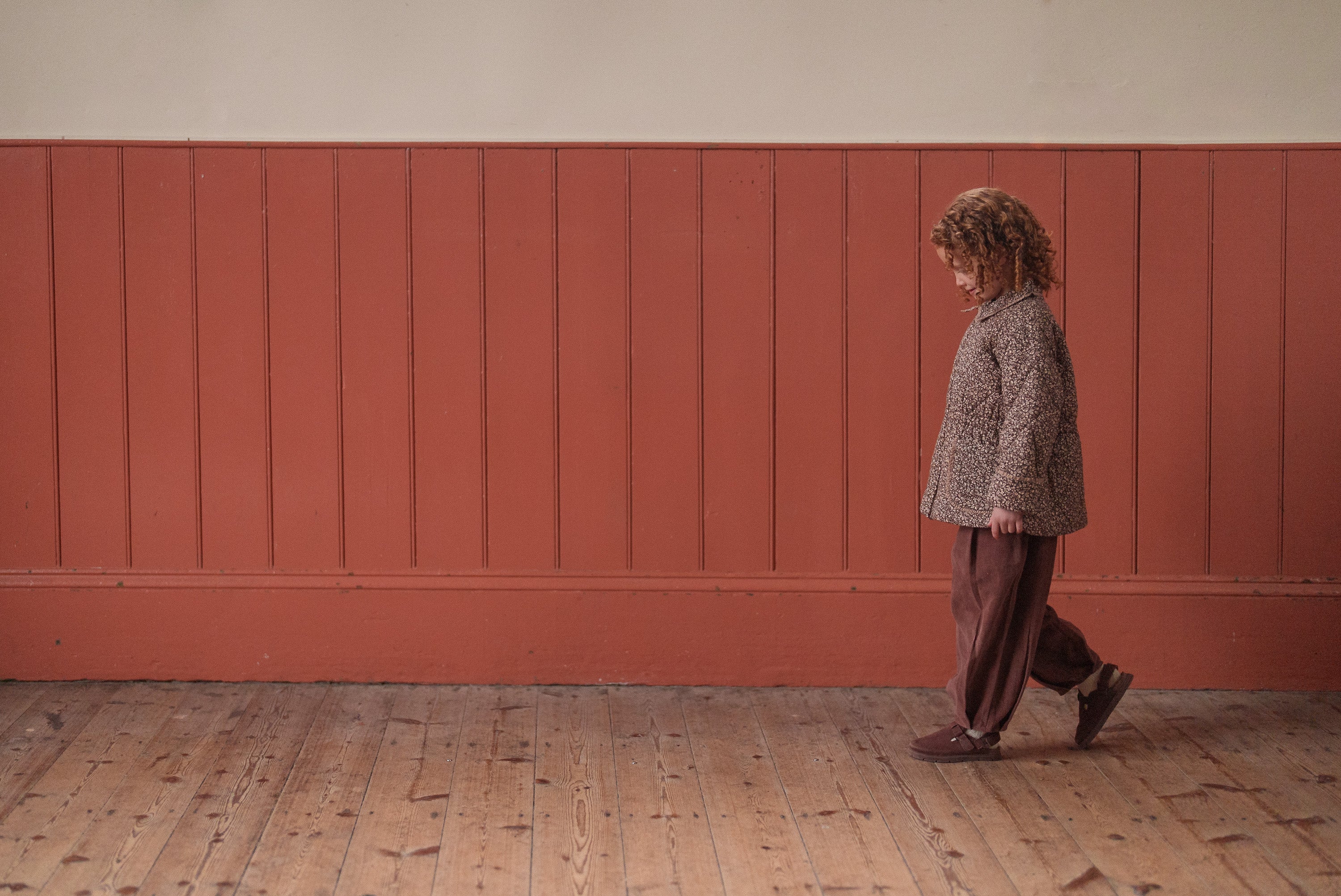 Young child wearing a brown and beige floral organic cotton jacket and matching pants, walking indoors on a wooden floor against a muted red paneled wall in vintage-style kids clothing.