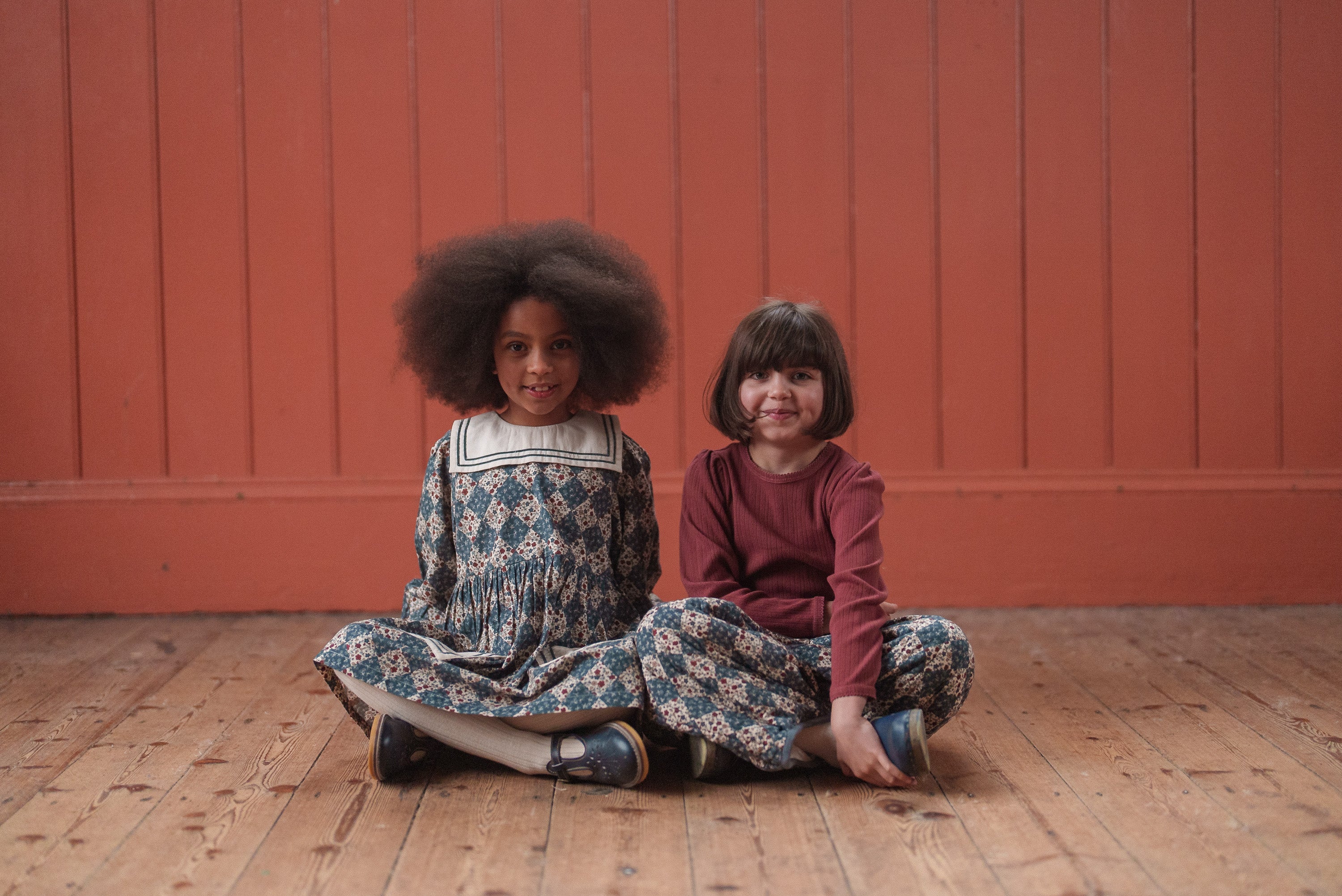 Two young girls sitting on a wooden floor wearing vintage-style, natural fabric children's clothing in a warm, cozy setting with a red background wall.