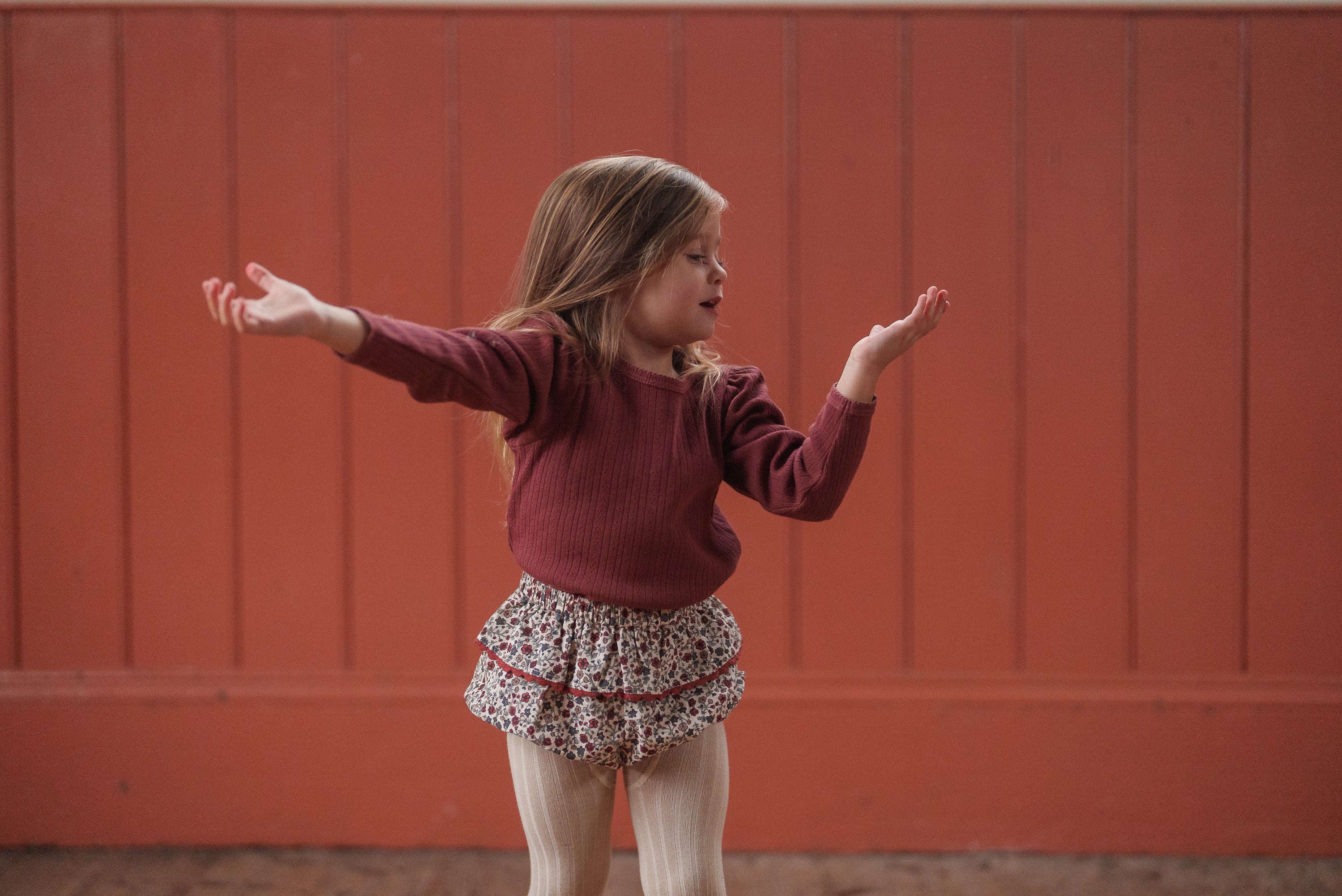 Young girl wearing a red organic cotton pointelle T-shirt and vintage style floral ruffle shorts with tights, standing indoors against a warm red wooden wall, showcasing sustainable kids clothing.