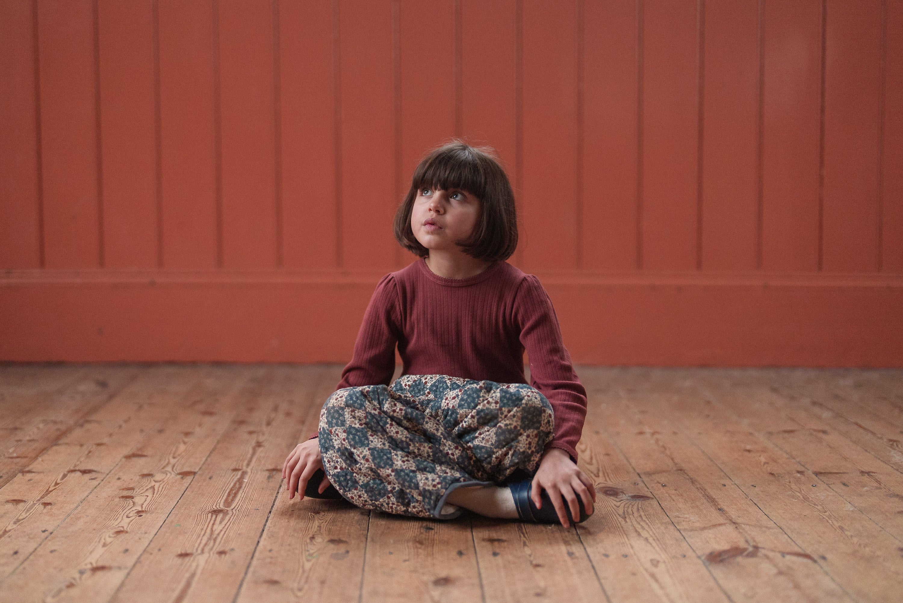 Young child sitting cross-legged on wooden floor wearing a red organic cotton long-sleeve top and vintage style patterned pants in a warm, natural setting