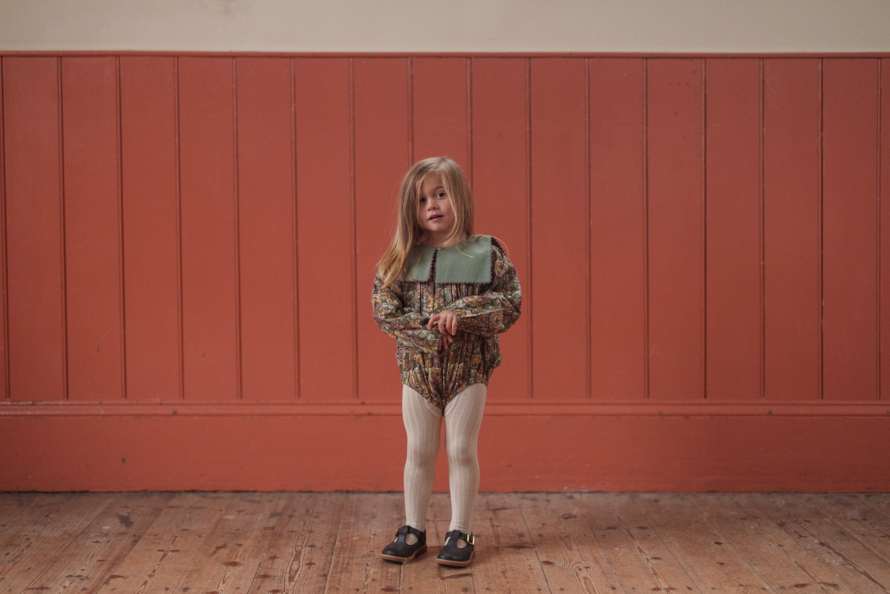 Young girl standing indoors wearing a green and pink floral romper made from organic cotton, styled with vintage-inspired tights and black shoes, embodying sustainable children's clothing.