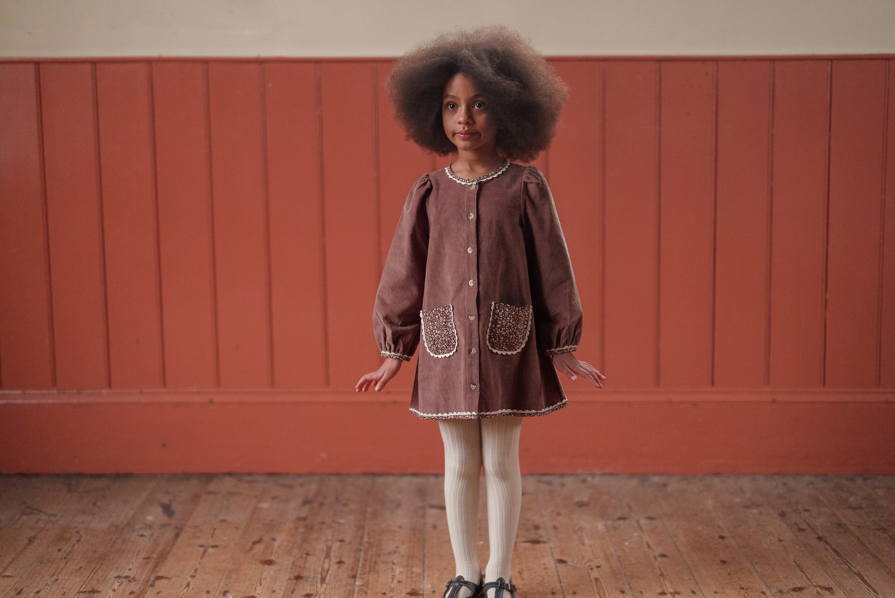 Young girl wearing a vintage style girls dress in nutmeg velvet with floral details, made from organic cotton, standing on a wooden floor against a red wall.