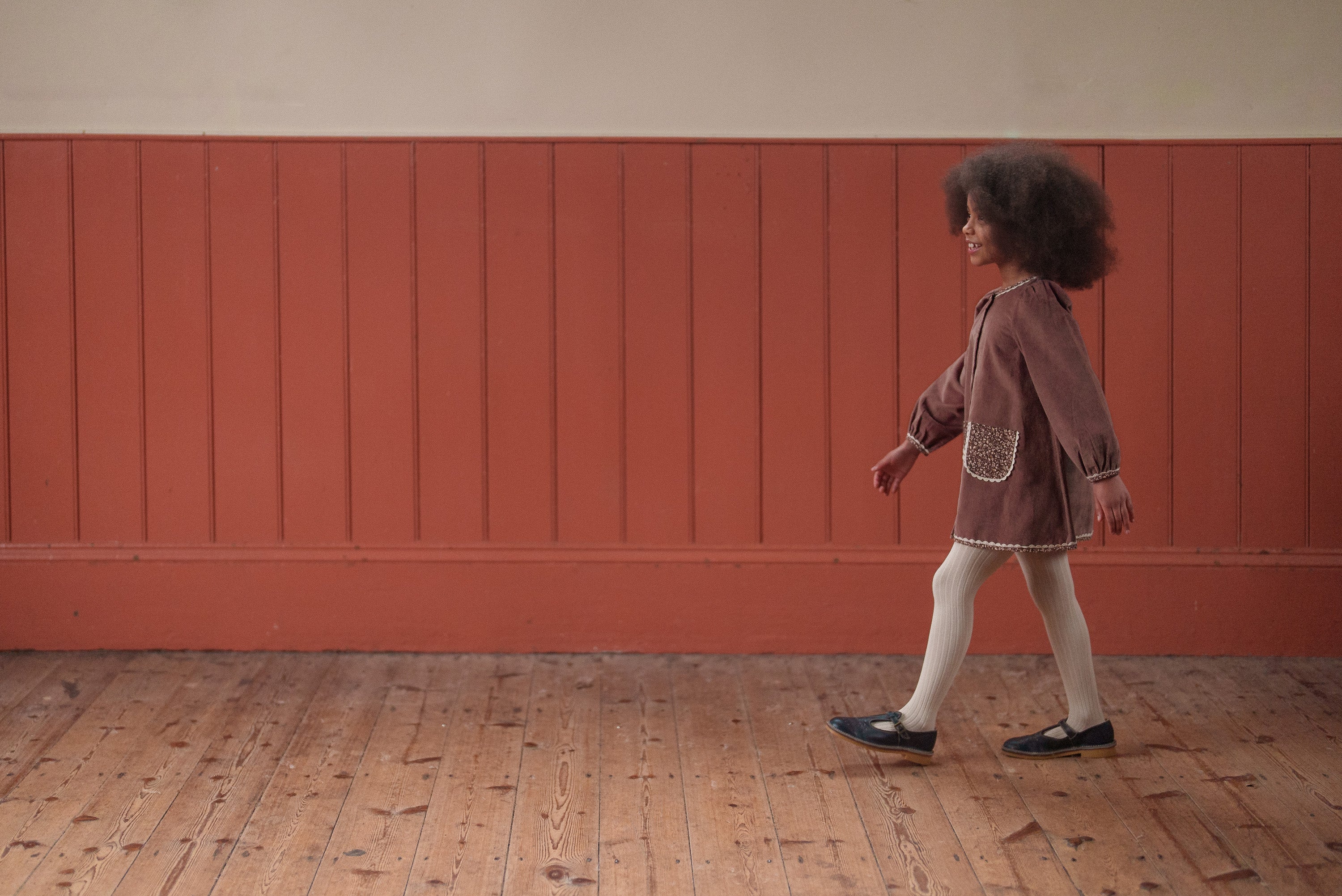 Young girl walking indoors wearing a vintage style dress in nutmeg velvet with organic cotton, paired with white tights and black shoes, showcasing sustainable kids clothing.