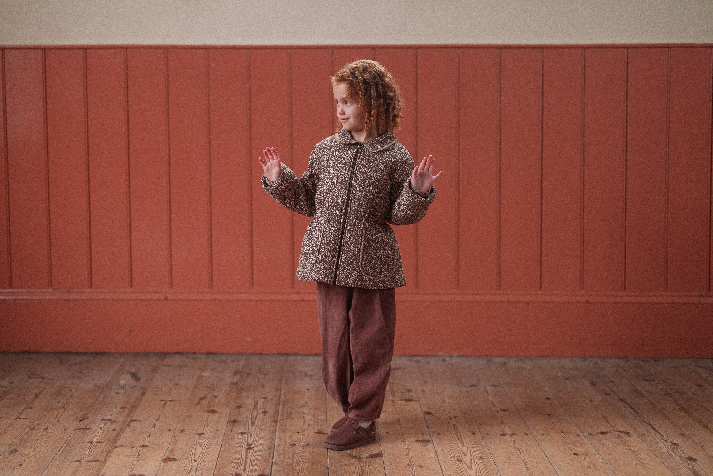 Young child with curly hair wearing vintage-style brown velvet trousers and a patterned jacket, standing on a wooden floor with a warm-toned wall background in a natural light setting.